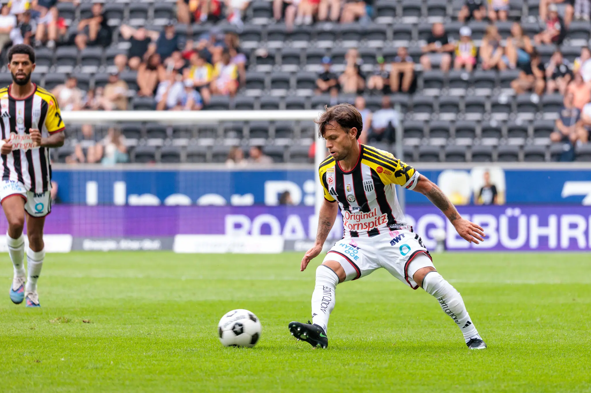 LASK-Spieler mit längeren Haaren im Anlauf bereitet einen Pass oder Schuss vor, im Hintergrund ein weiterer LASK-Spieler und Zuschauer.