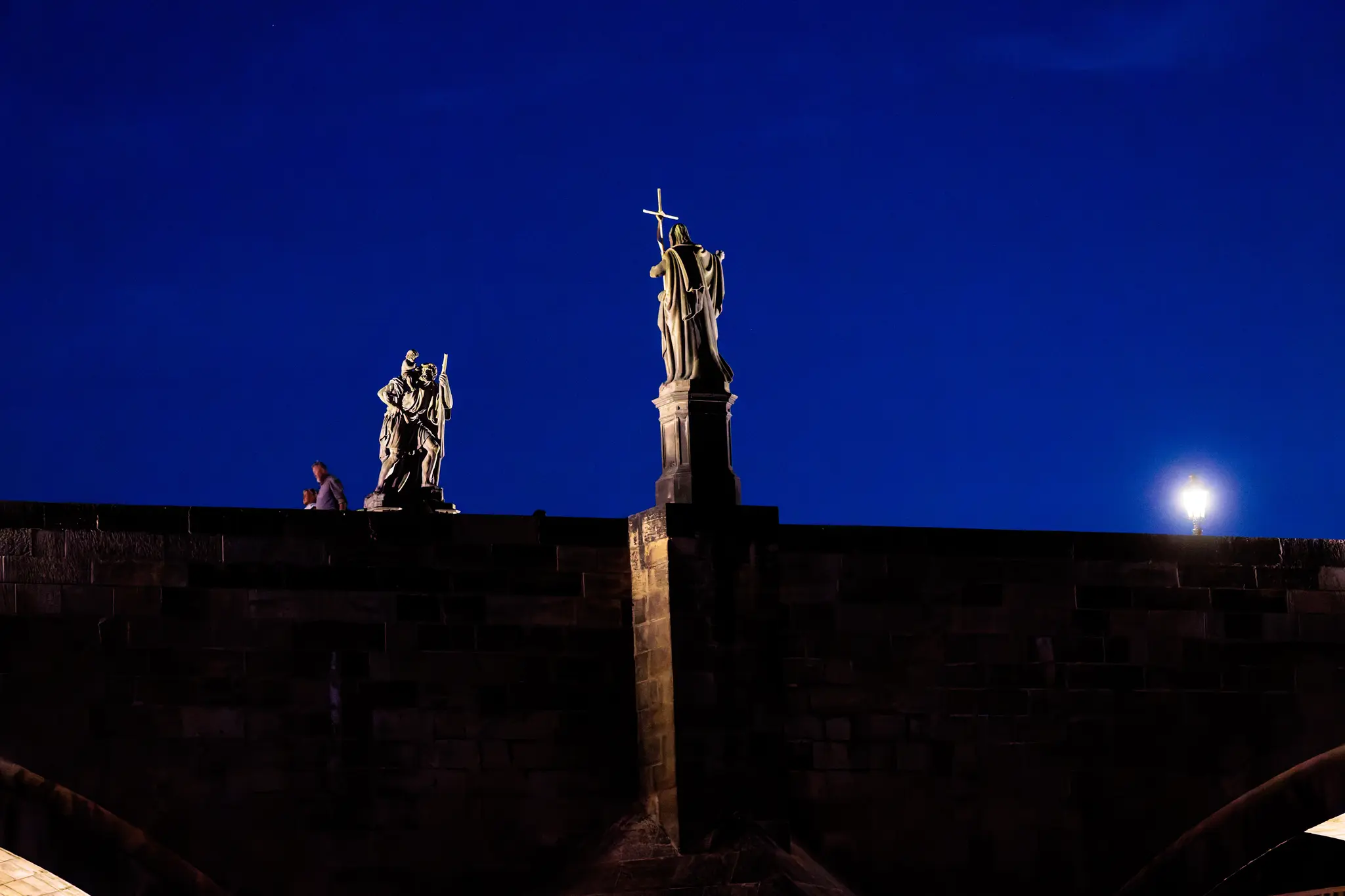 Barockstatuen auf der Karlsbrücke heben sich vor dem tiefblauen Nachthimmel ab; ein Spaziergänger lehnt am Geländer neben einer leuchtenden Laterne.
