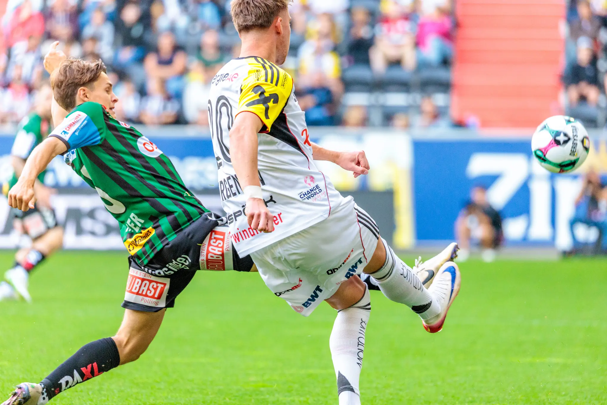Ein Spieler des LASK in weißem Trikot flankt den Ball nach vorne, während ein SV Ried-Spieler in grün-schwarzem Trikot versucht, die Aktion zu stören
