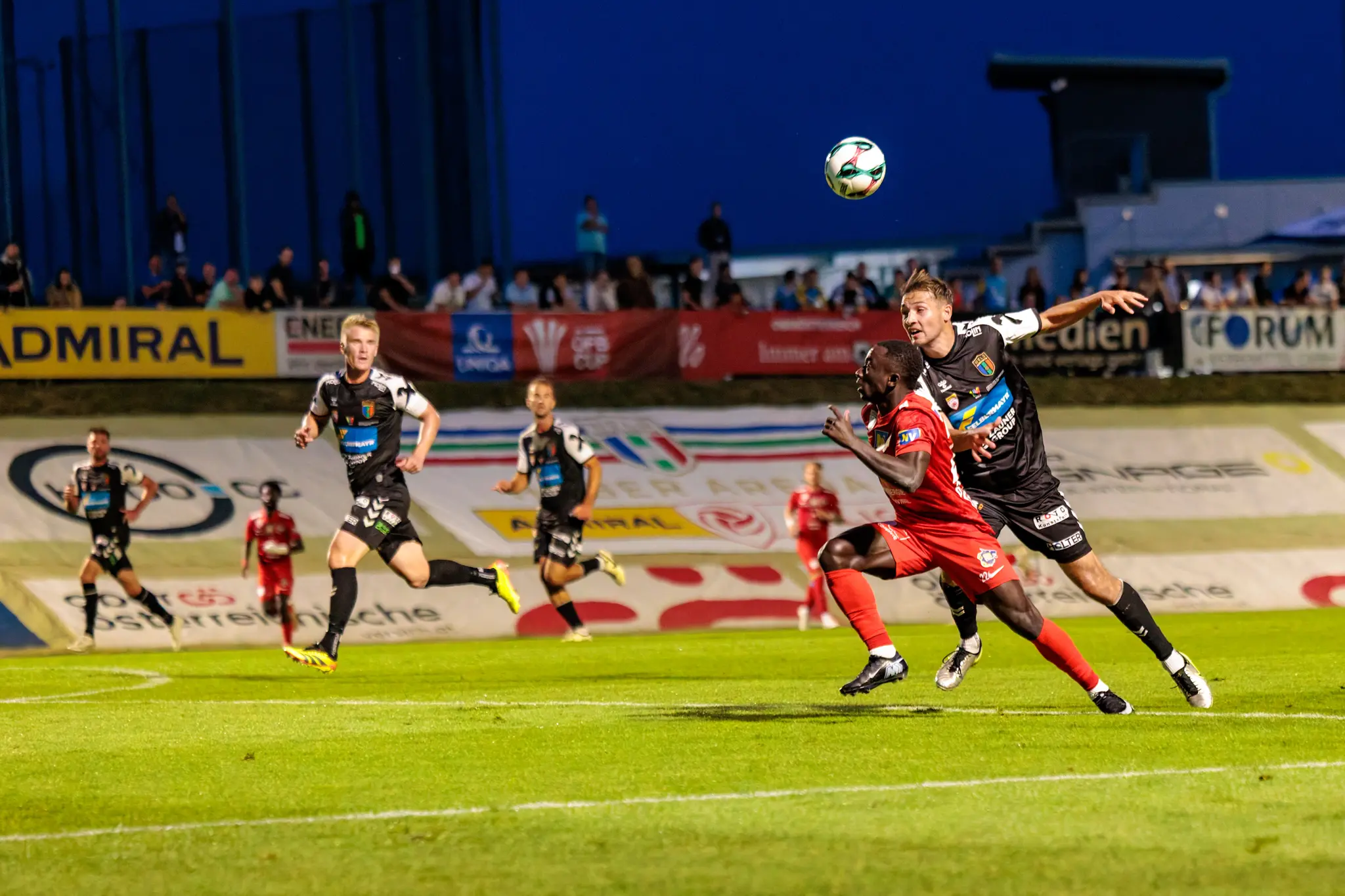 Zwei Fußballspieler, einer in rotem und einer in schwarzem Trikot, duellieren sich im Strafraum um einen hoch in der Luft schwebenden Ball während eines ÖFB-Cup-Spiels