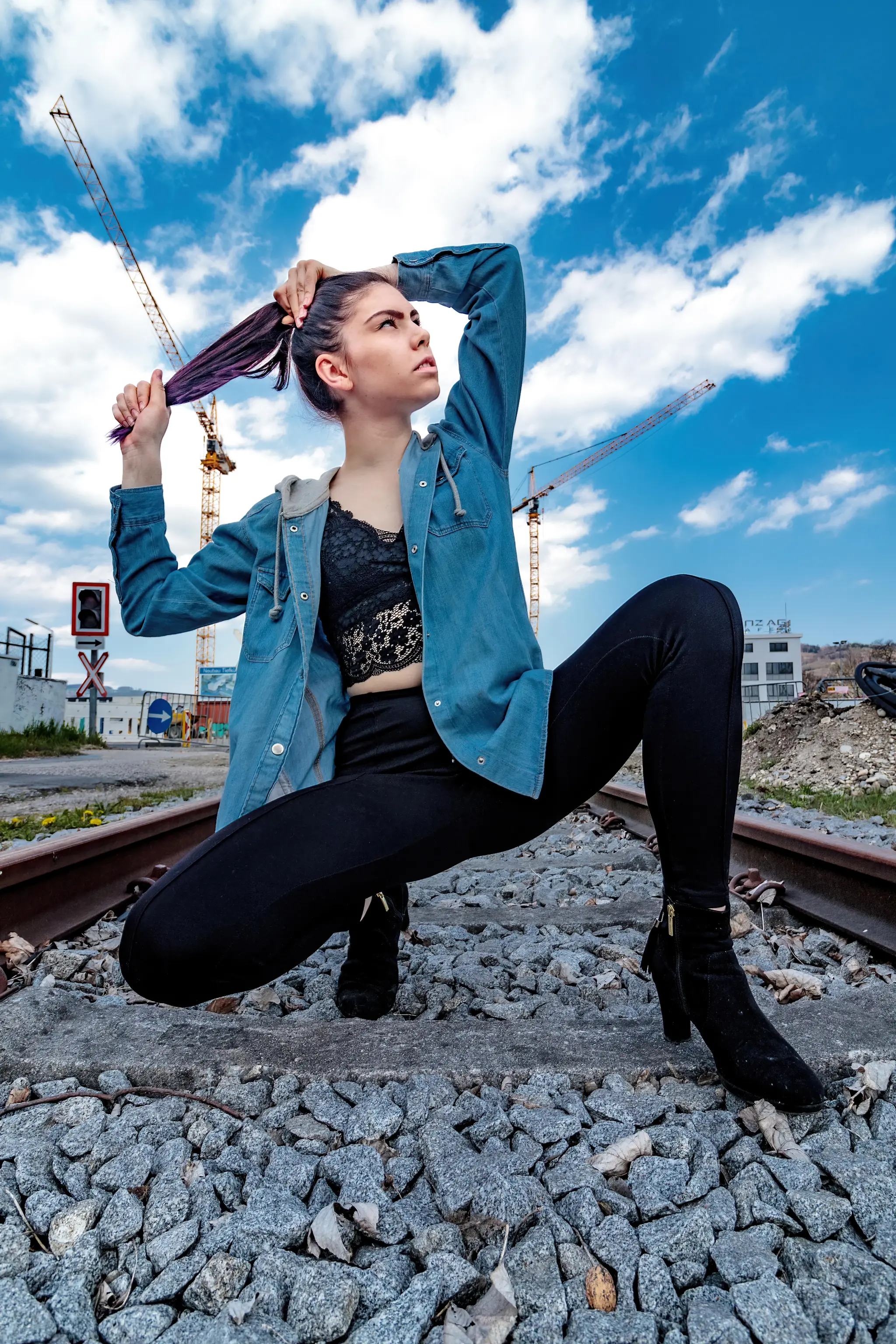 Eine junge Frau in blauer Jeansjacke hockt auf Bahnschotter, richtet ihr Haar und blickt unter einem weiten blauen Himmel mit Baukränen nach oben