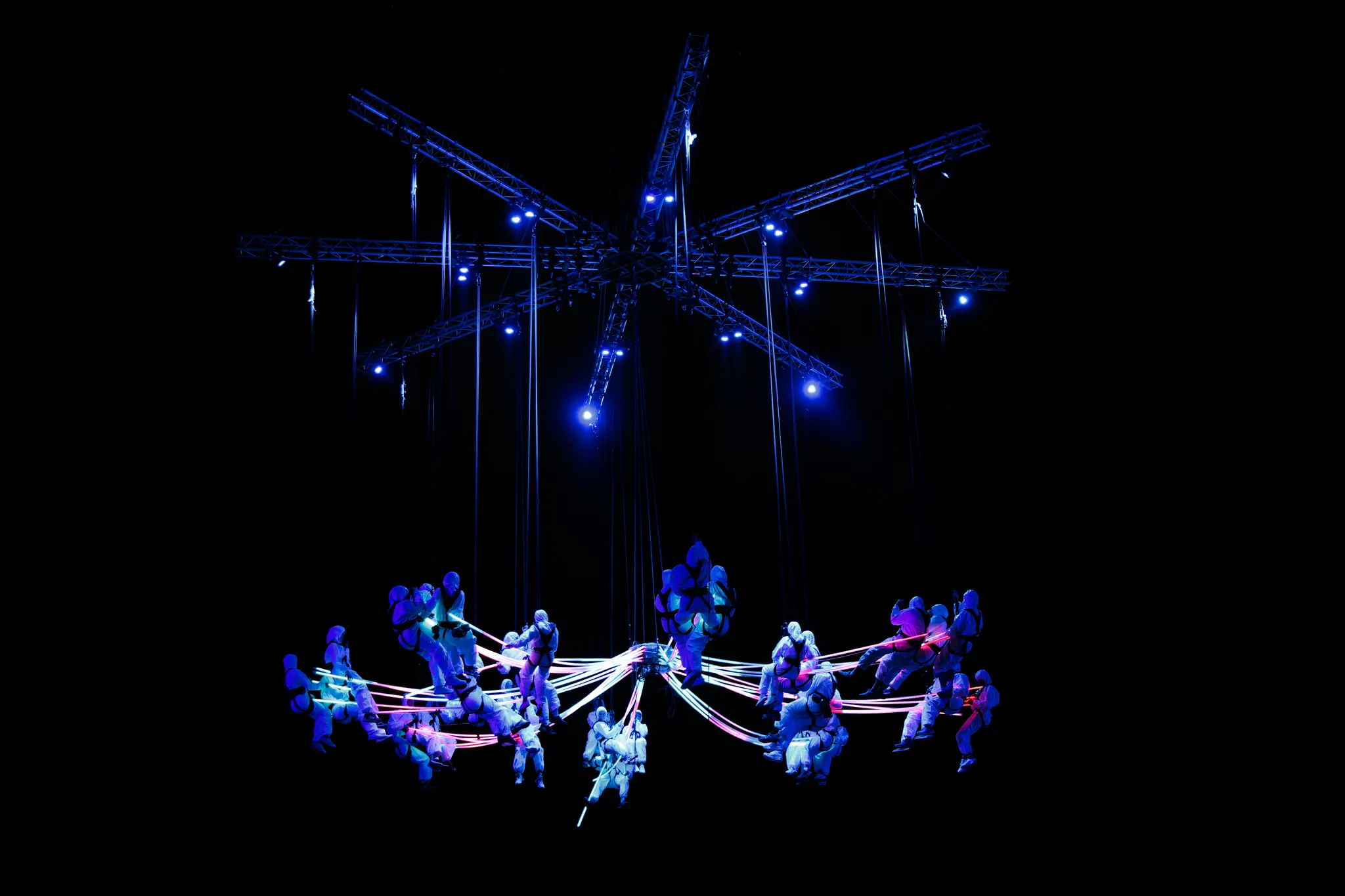 Eine dynamische Lichtshow im Rahmen der Linzer Klangwolke 2024 mit schwebenden Akrobaten in leuchtenden Anzügen, die durch strahlende Linien verbunden sind, unter einer blau beleuchteten Metallkonstruktion vor dunklem Hintergrund