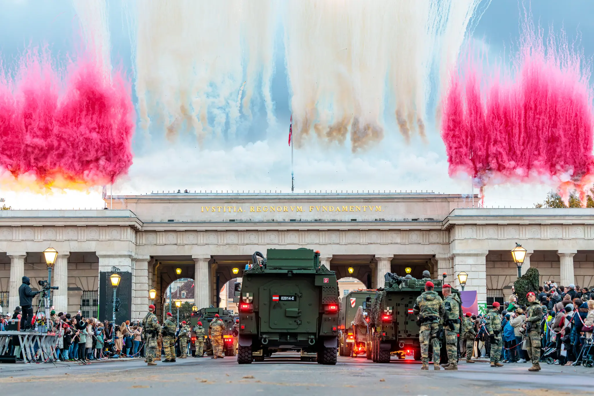 Über dem Äußeren Burgtor steigen zum Nationalfeiertag rote und weiße Rauchsäulen in den Himmel, davor rollen Militärfahrzeuge mit Soldaten.
