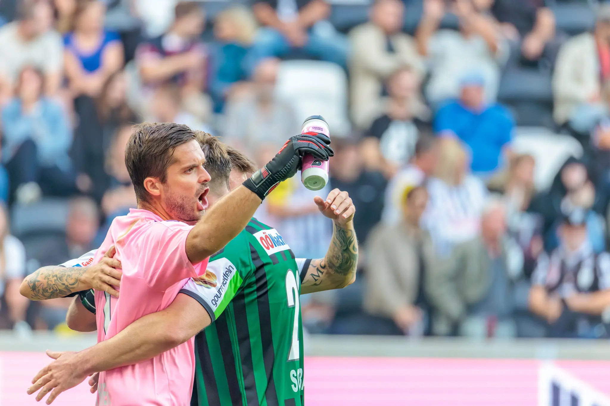 Der Torhüter des SV Ried in pinkem Trikot und Handschuhen jubelt mit einem Mitspieler in grün-schwarz gestreiftem Trikot über den Derby-Sieg gegen den LASK Linz