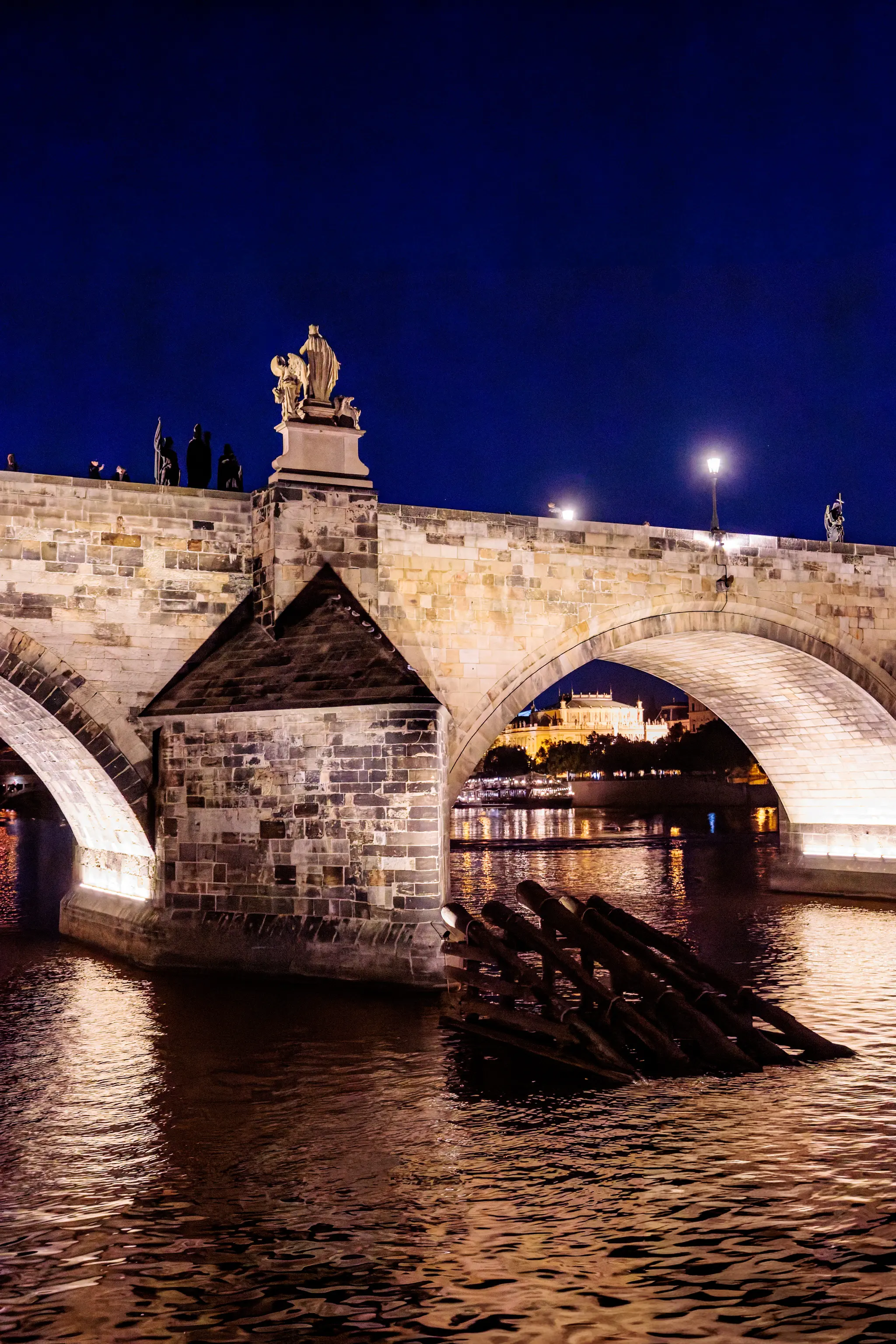 Durch den Bogen der Karlsbrücke leuchtet das Nationaltheater über die Moldau; Eisbrecher-Pfähle und Wasserreflexionen tragen die warmen Stadtlichter.