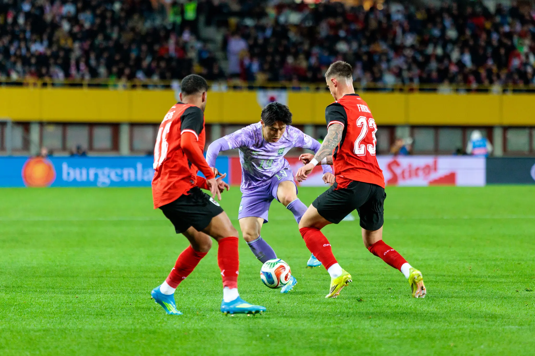 Ein südkoreanischer Nationalspieler im lila Trikot behauptet im Zweikampf den Ball bei einem Dribbling gegen zwei österreichische Nationalspieler in rot-schwarzer Spielkleidung