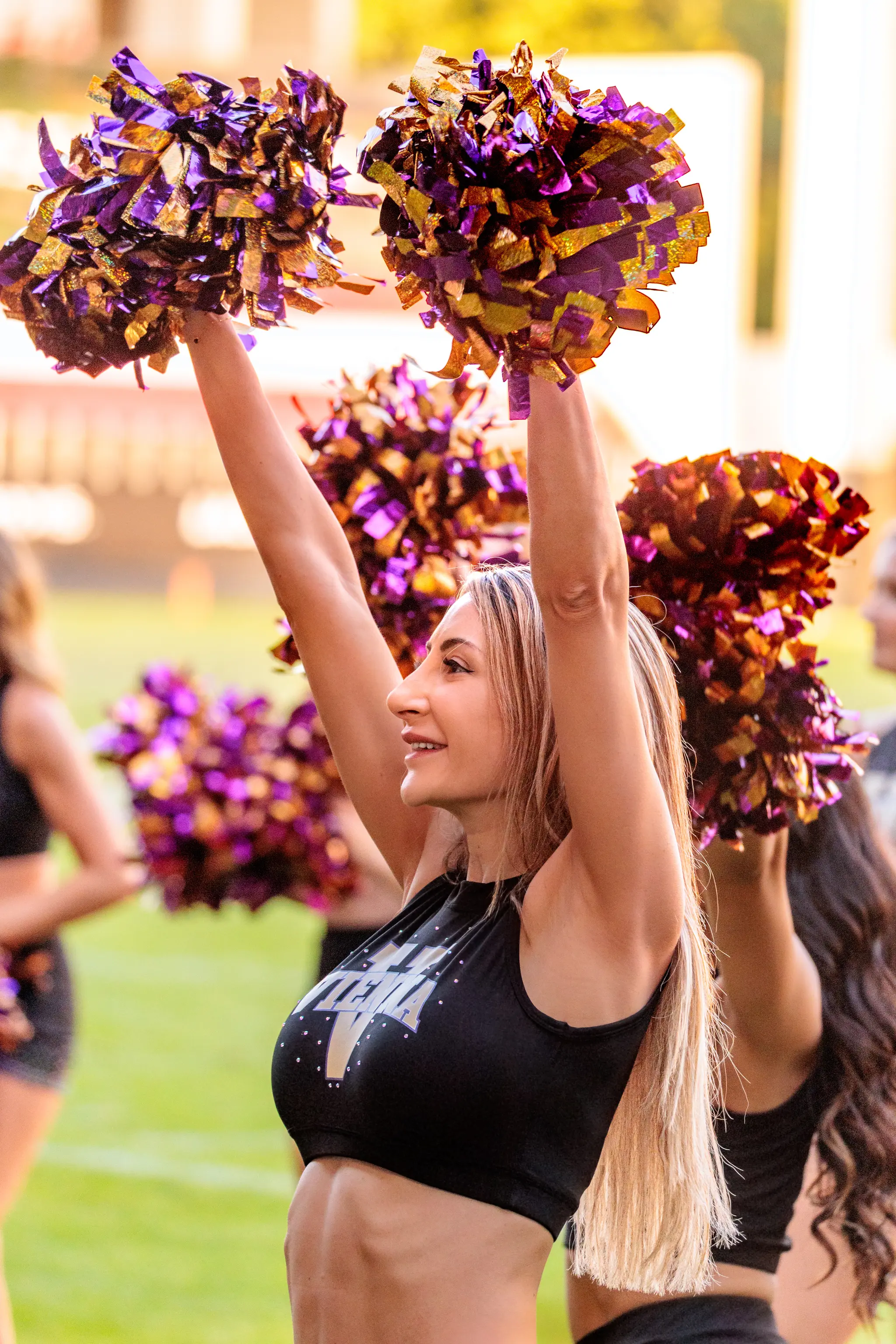 Cheerleaderin des Spirit Squad hebt beide Arme mit violett-goldenen Pompons in die Höhe.