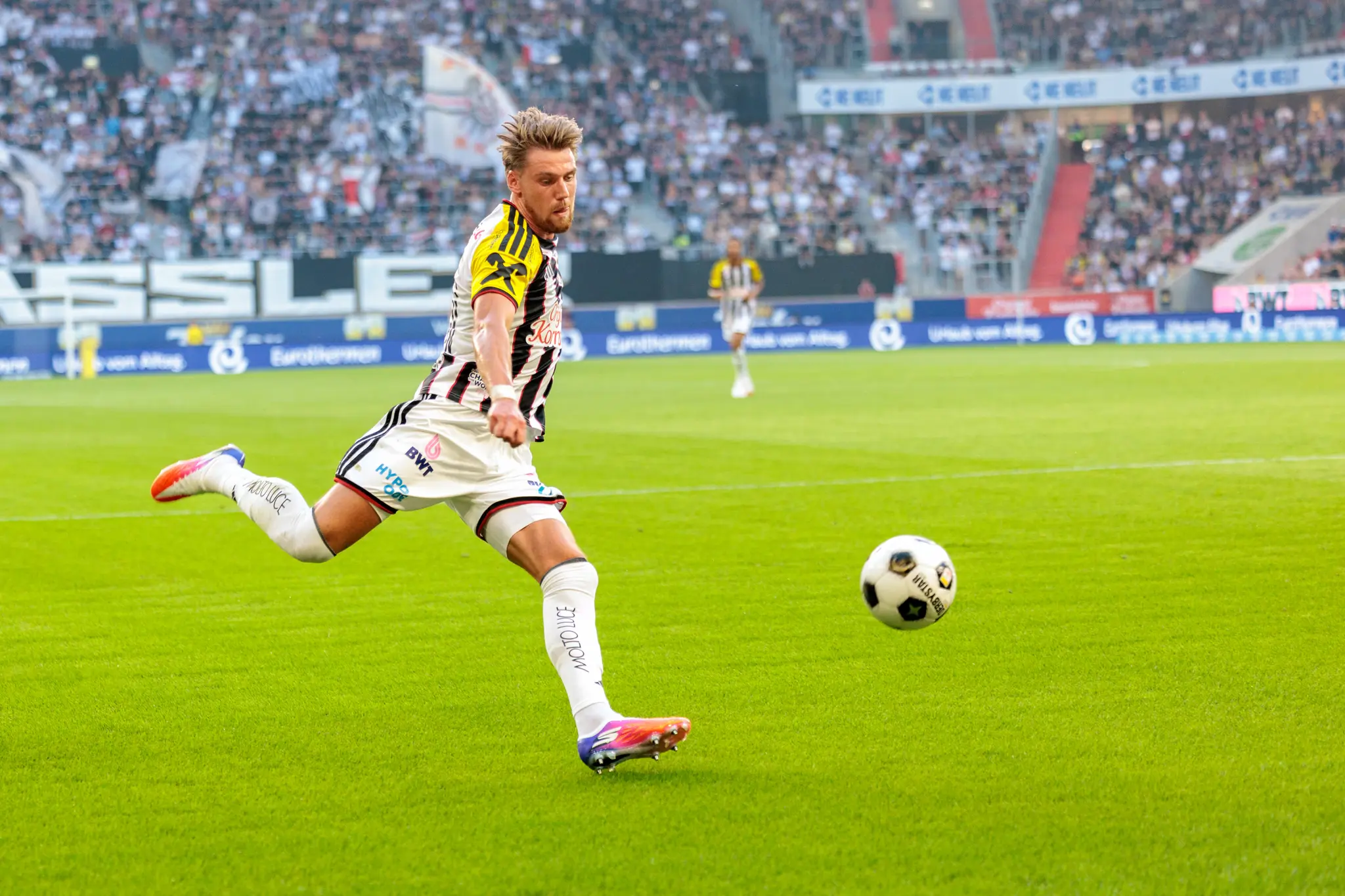 LASK-Spieler schießt den Ball mit dem rechten Fuß Richtung Tor, während der Ball in der Luft schwebt und die Abwehr reagiert