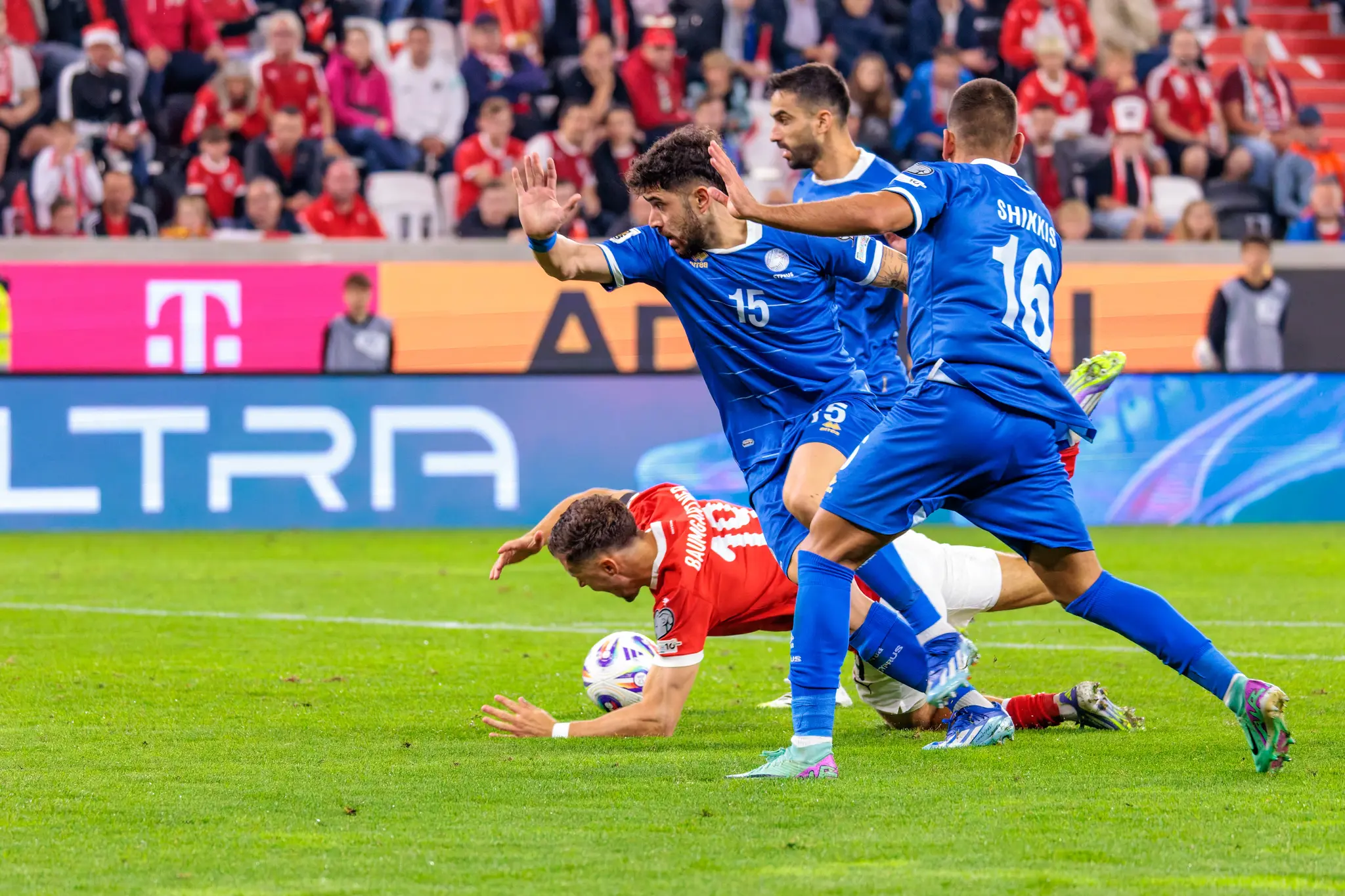 Der österreichische Spieler Christoph Baumgartner im roten Trikot ist nach einem Zweikampf mit zwei zypriotischen Gegenspielern im blauen Trikot zu Boden gegangen