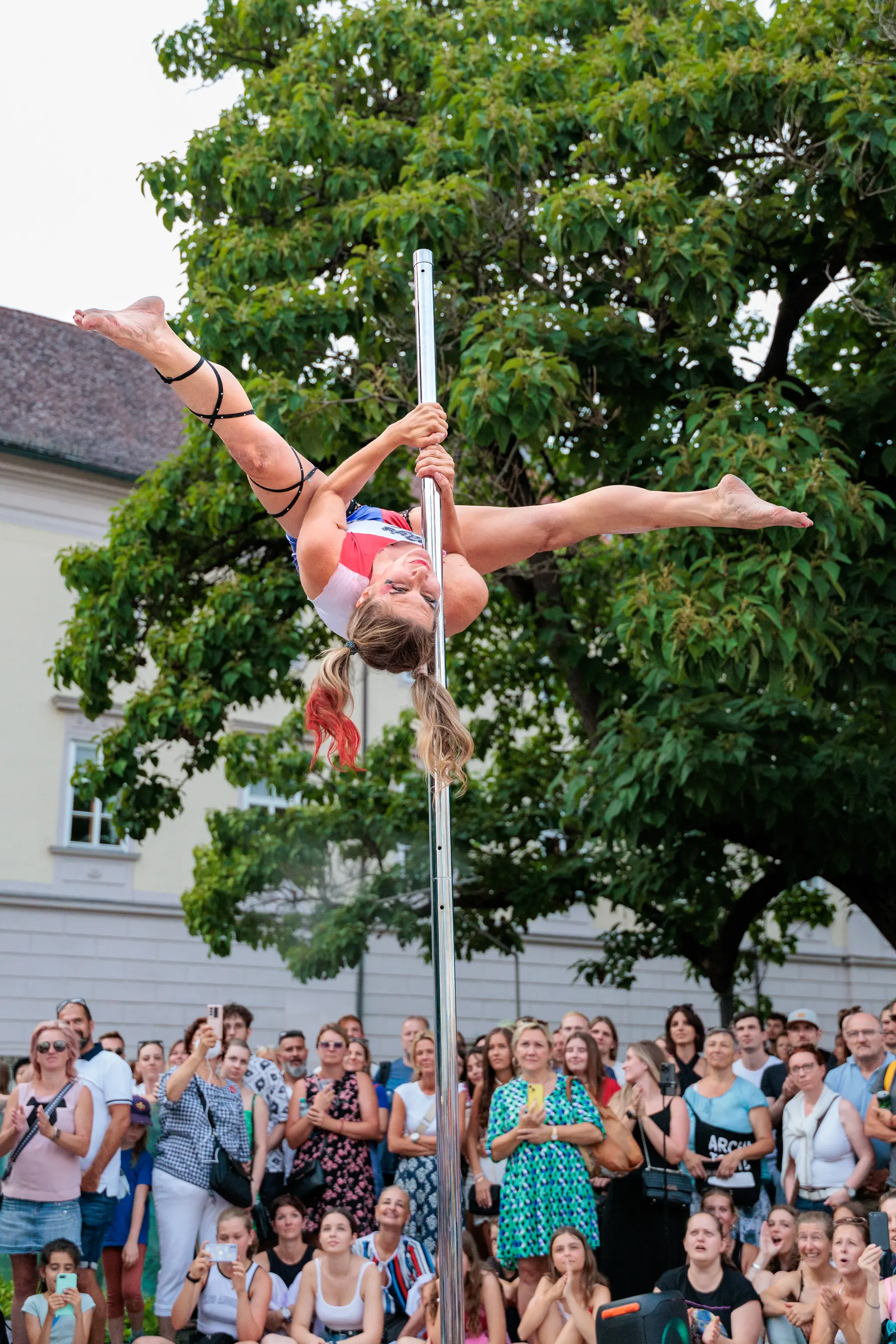 Eine Frau in Poledance-Sportkleidung vollführt kopfüber eine Spagat-Pose an einer Stange bei einer Outdoor-Vorstellung vor dem Linzer Landhaus während des Pflasterspektakels 2024 