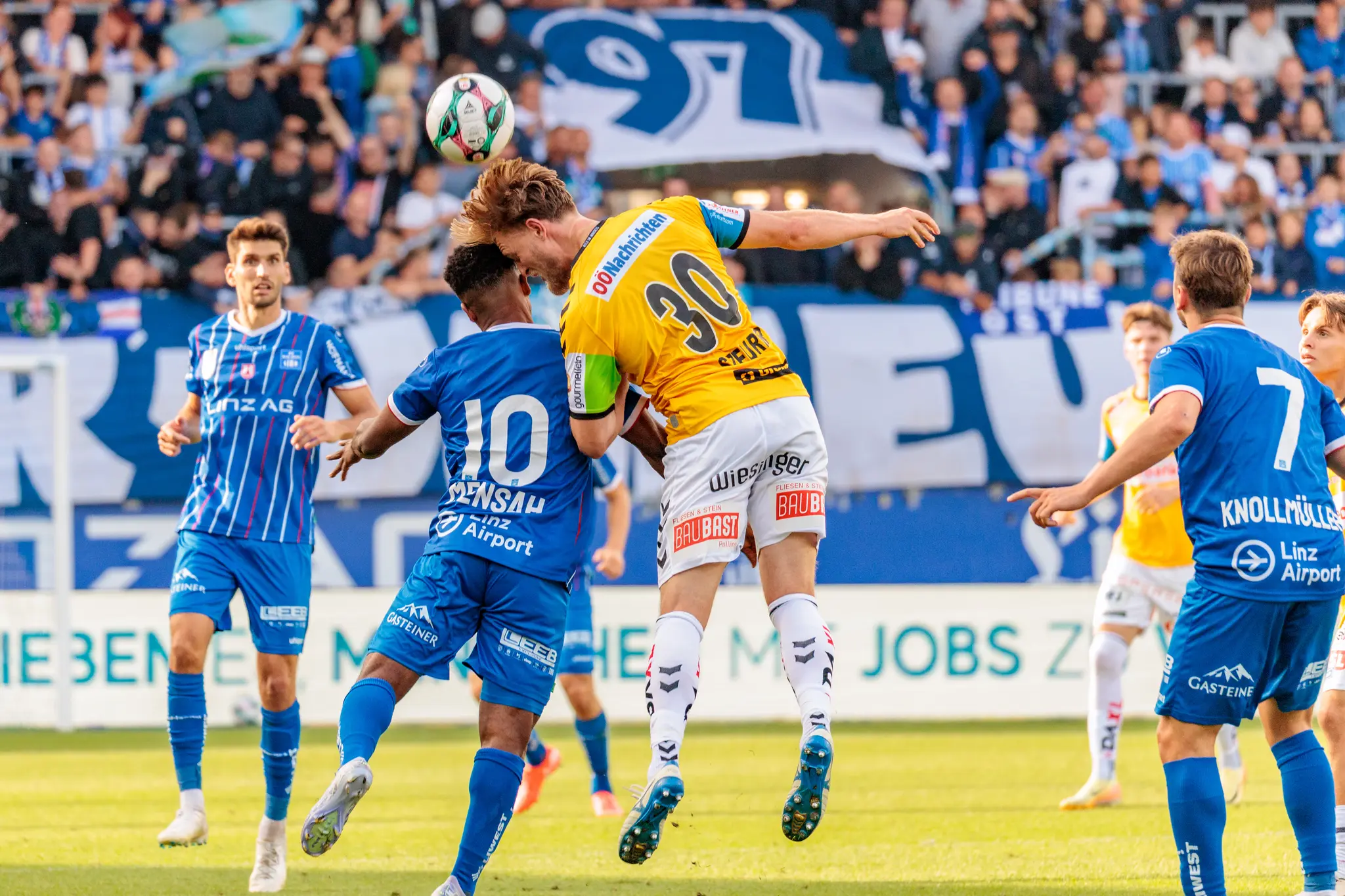 Ein Fußballer in gelbem Trikot und ein Spieler in blauem Trikot springen beim Kopfballduell um den Ball, während im Hintergrund Zuschauer auf den Tribünen zu sehen sind