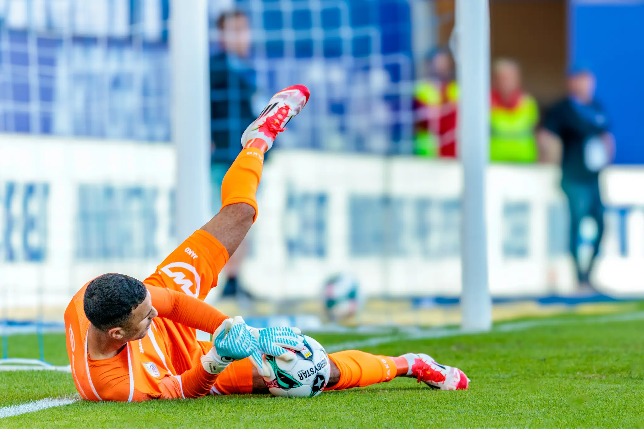 Der Tormann des SCR Altach, Dejan Stojanovic, zeigt in seinem leuchtend orangen Trikot eine spektakuläre Parade, bei der er sich voll ausstreckt und den Ball sicher am Boden festhält