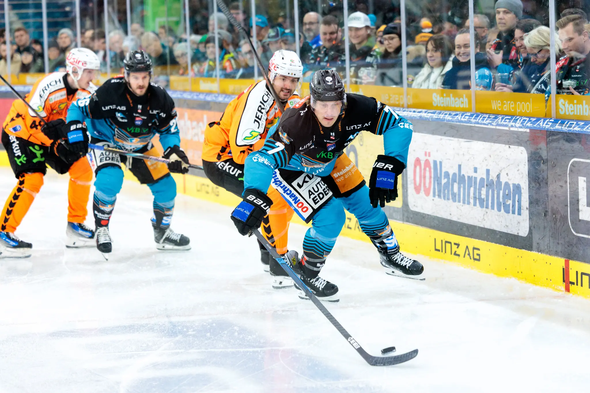 Ein Eishockeyspieler der Black Wings Linz im Angriff mit dem Puck auf dem Eis, dicht gefolgt von einem Spieler der Grazer 99ers