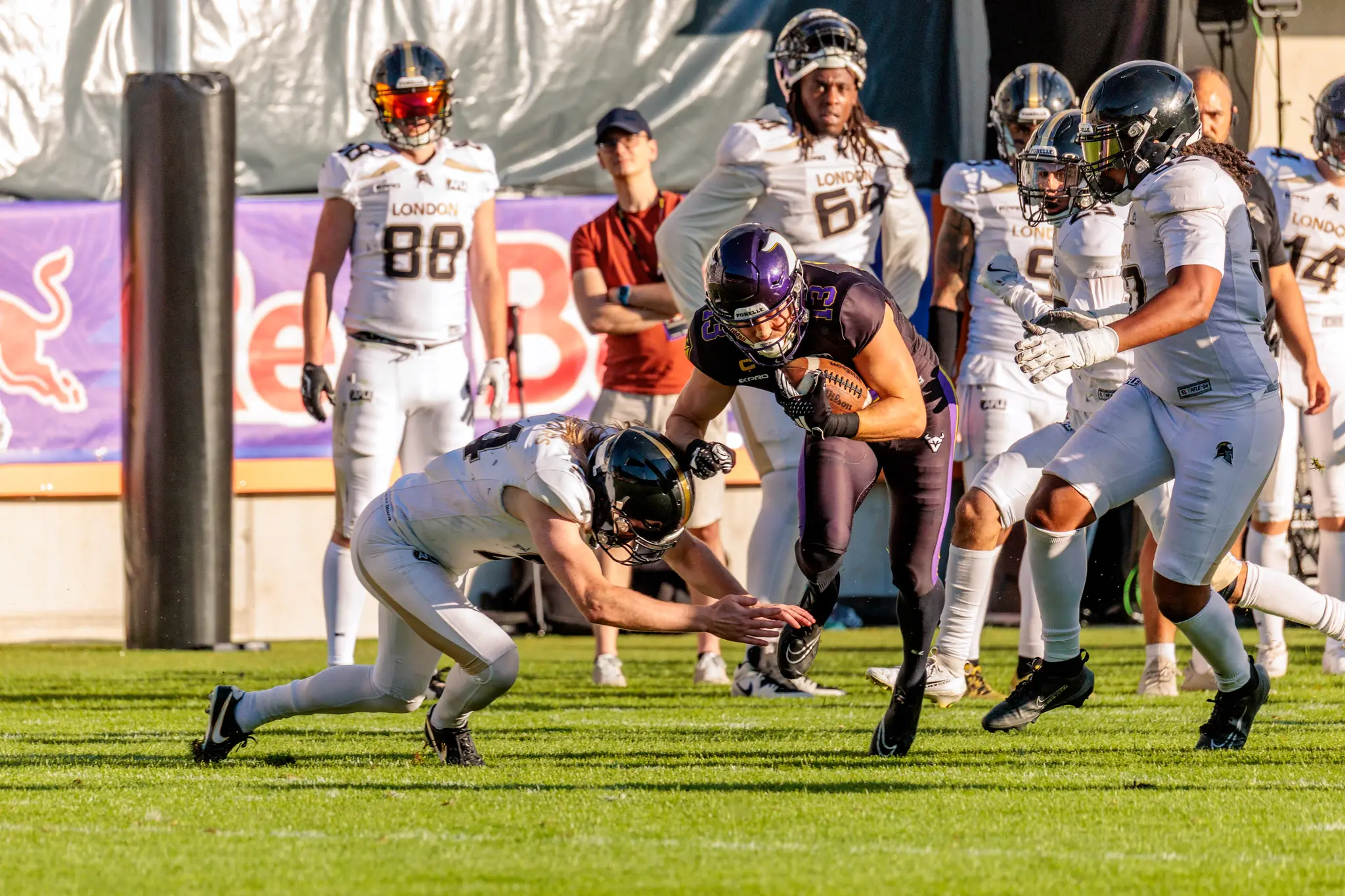 Philipp Haun (13) der Vienna Vikings trägt den Ball im ersten Viertel, während ein London-Verteidiger mit voller Länge zu einem Hecht-Tackle ansetzt.