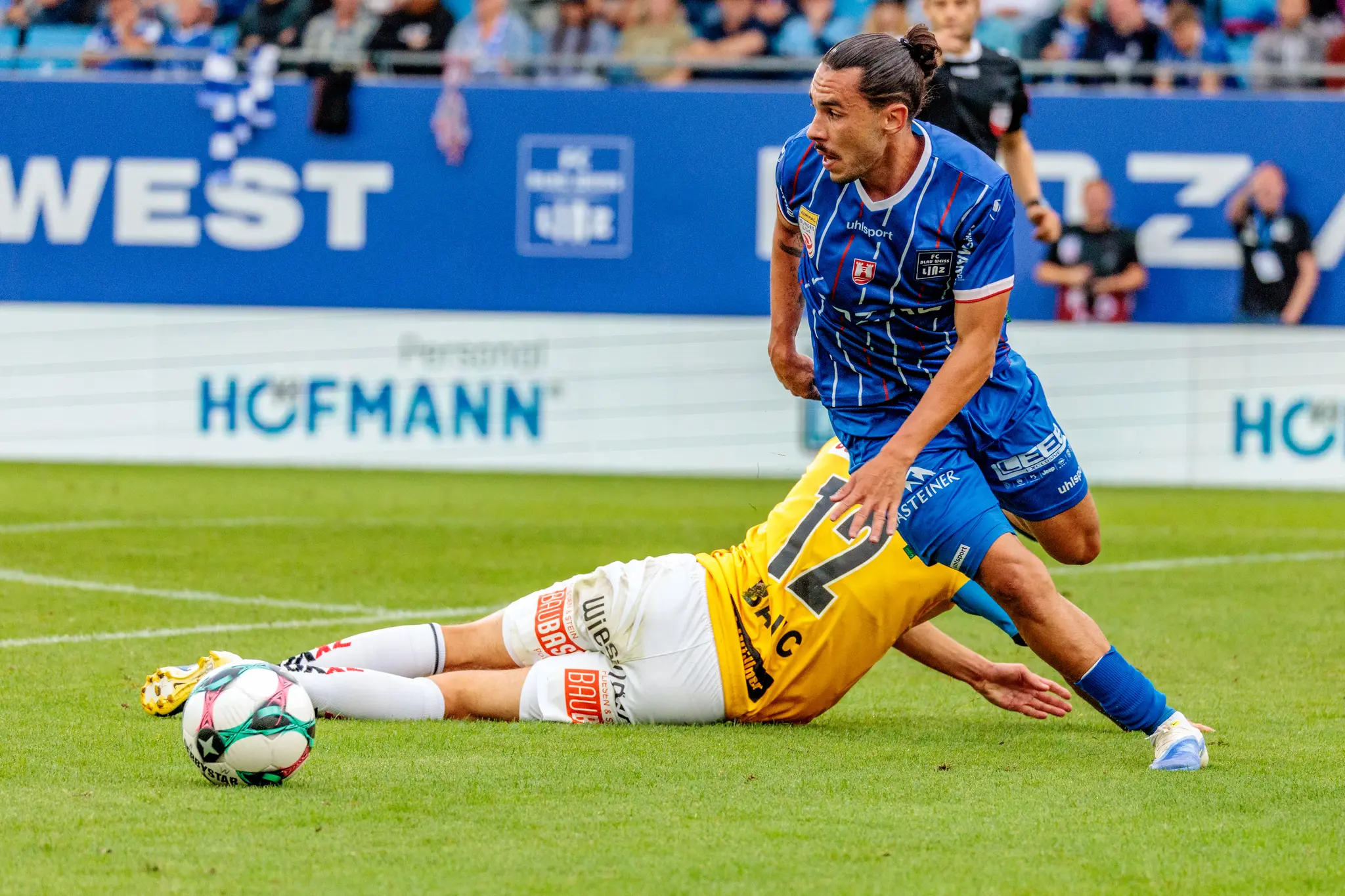 Ein Spieler von Blau-Weiß Linz steht über einem am Boden liegenden Gegner von SV Ried nach einem intensiven Zweikampf, der Fußball liegt in der Nähe