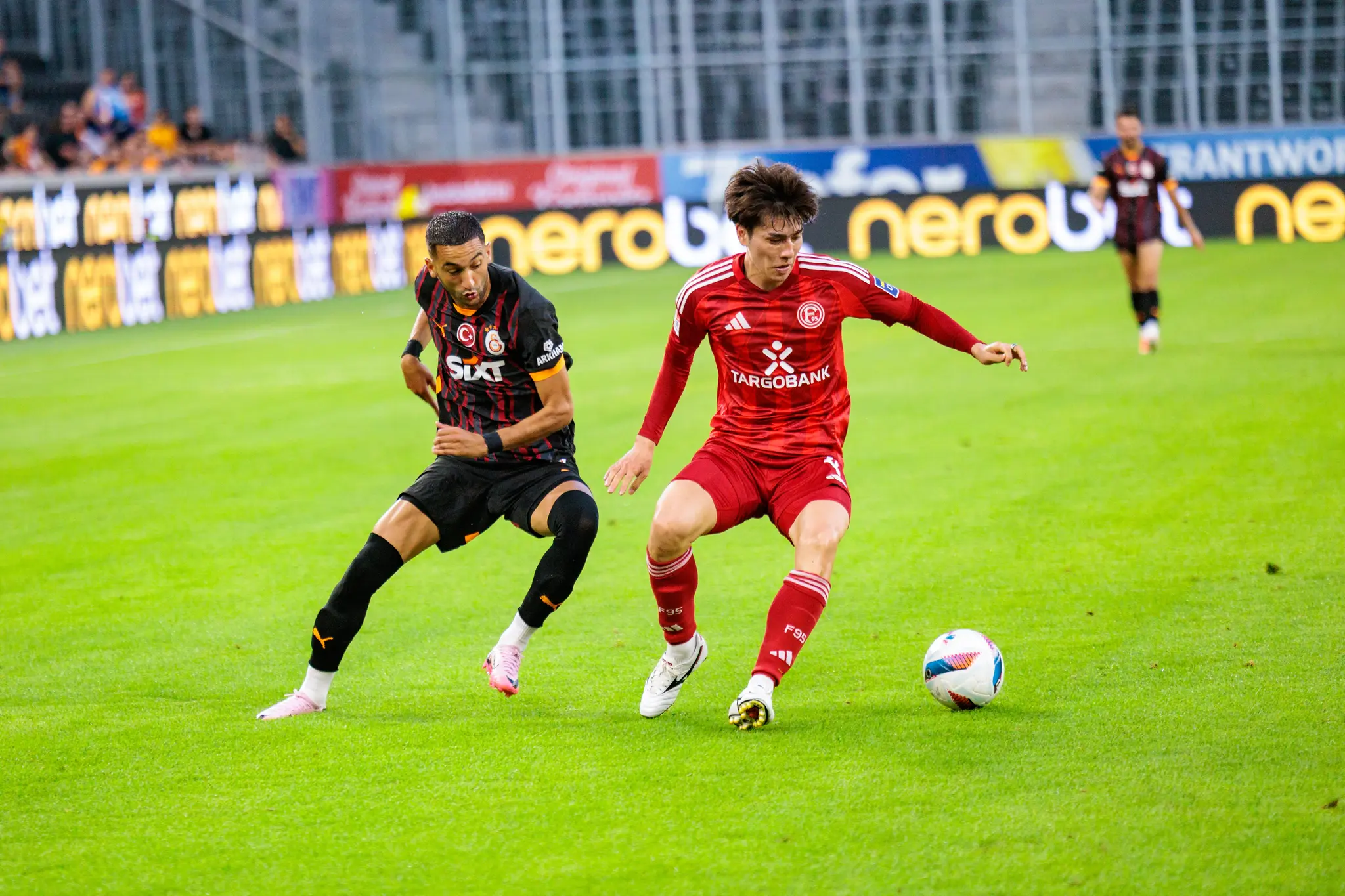 Zwei Fußballspieler in roten und dunklen Trikots duellieren sich auf dem grünen Rasen um den Ball 