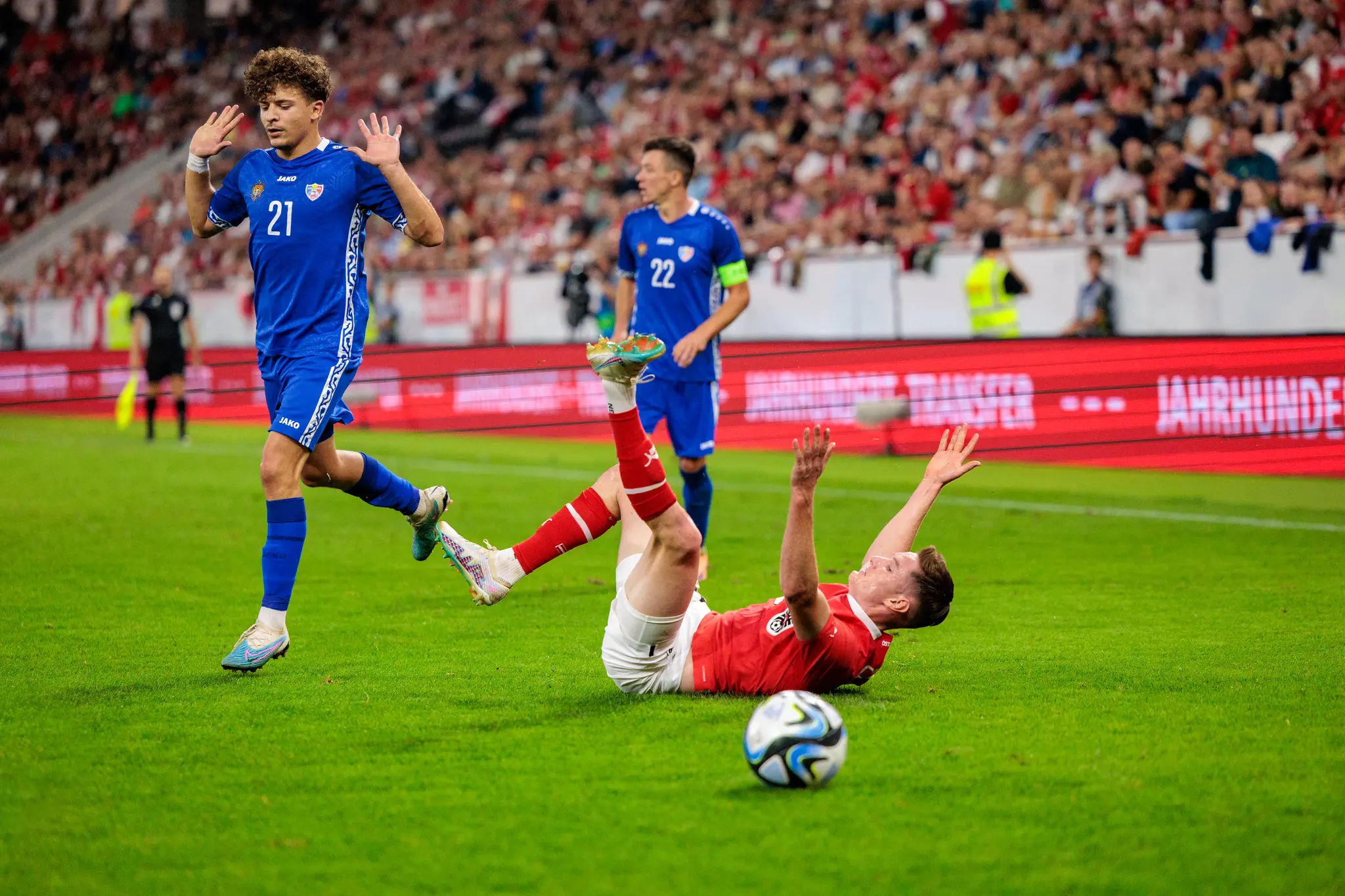 Aktionsszene aus einem Fußballspiel, bei der ein Spieler in blauem Trikot die Hände hebt, während ein Gegenspieler in rotem Trikot auf dem Boden liegt