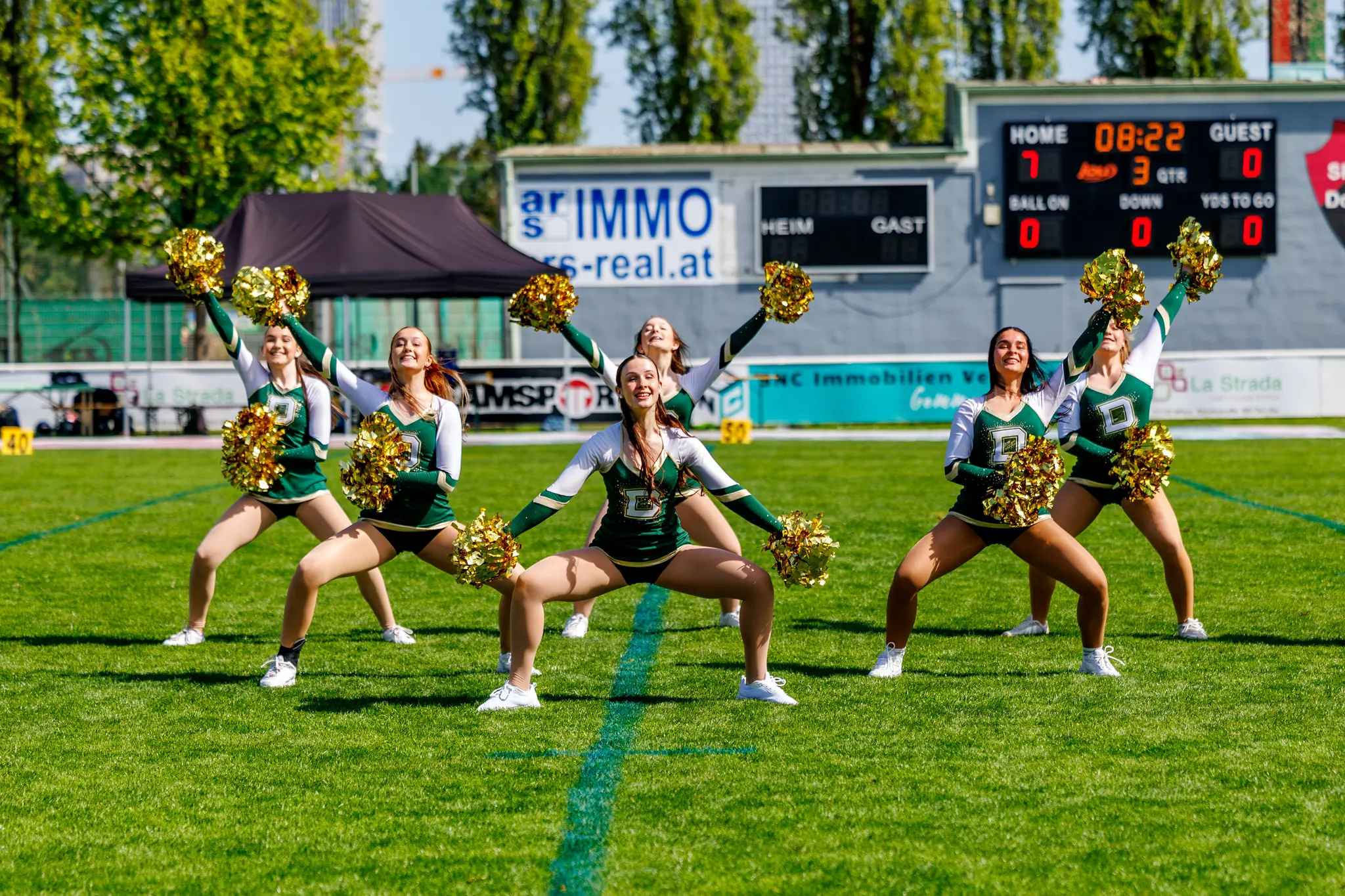 Sechs Cheerleader der Premium Dancers in grün-weißen Uniformen präsentieren eine dynamische Tanzpose mit goldenen Pompons