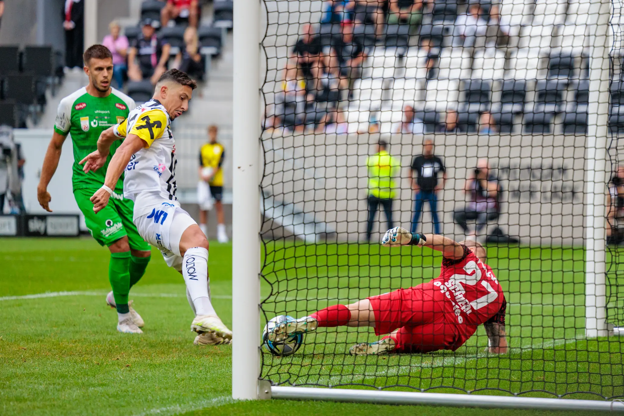Ein LASK-Fußballspieler in hellem Trikot schießt den Ball auf das Tor, während der gegnerische Torhüter in rotem Trikot abtaucht, um ihn abzuwehren