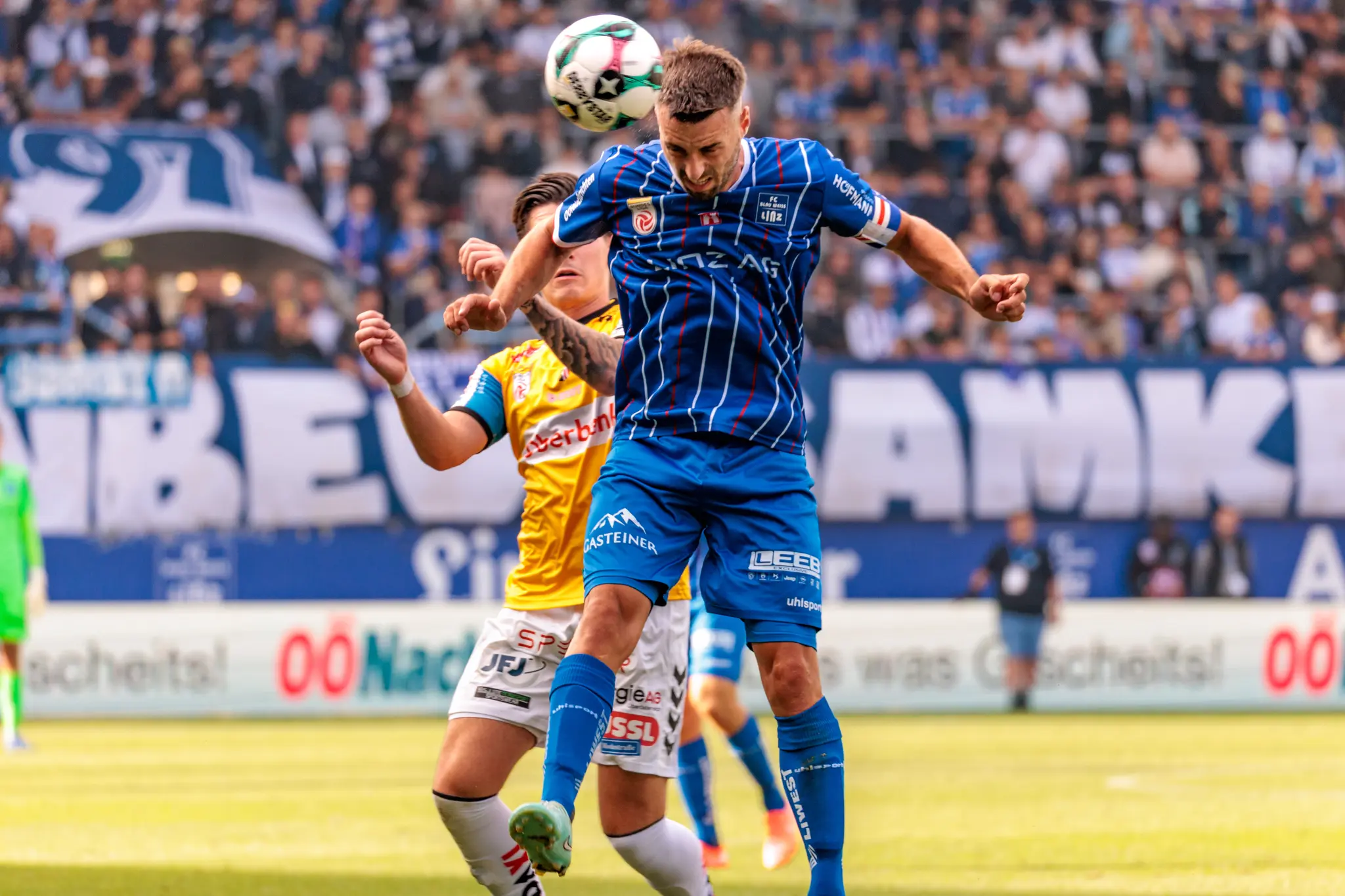 Ein Spieler des FC Blau-Weiß Linz im blauen Trikot und ein Spieler des SV Ried im gelben Trikot liefern sich ein dynamisches Kopfballduell um den Fußball