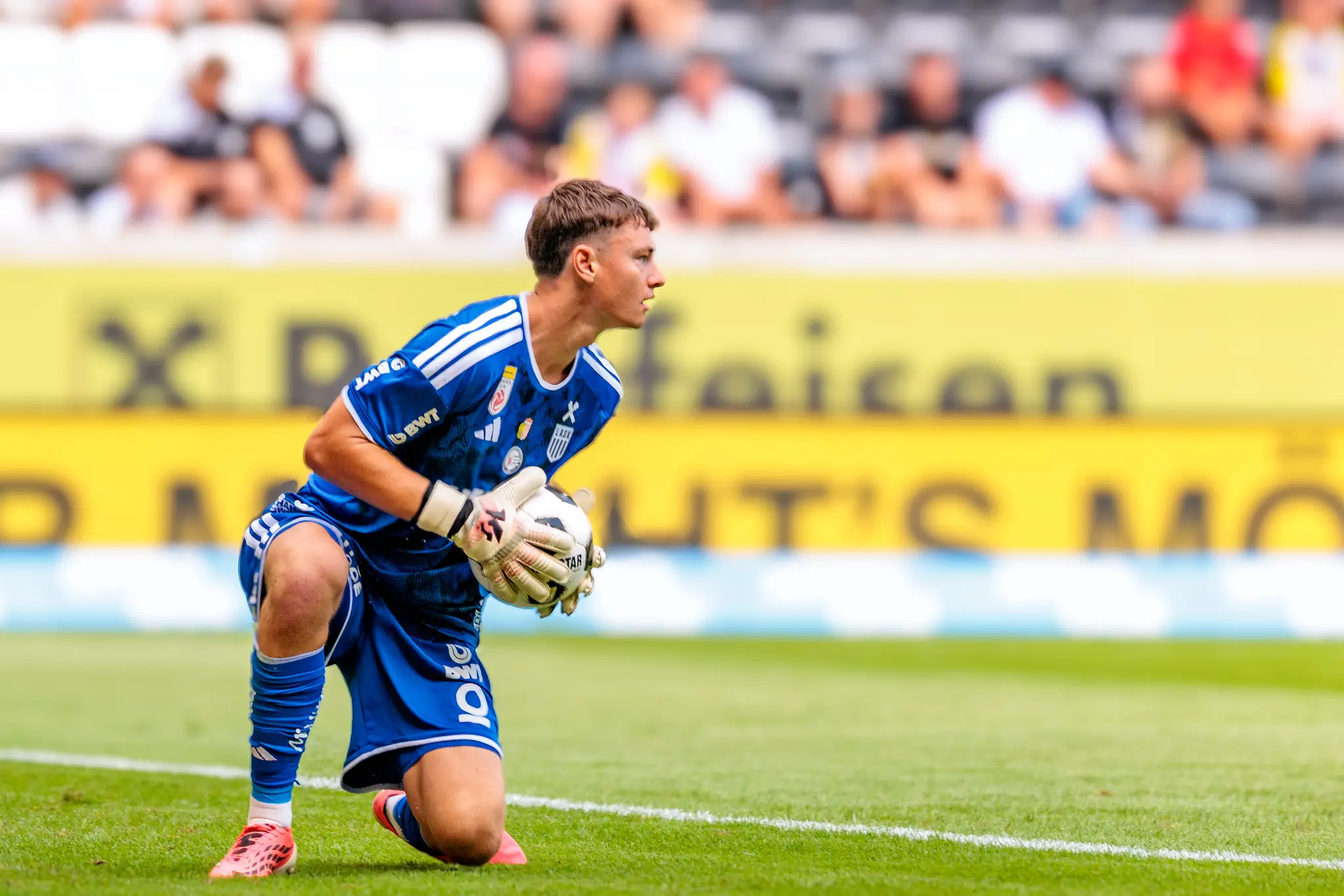 LASK-Tormann Jungwirth kniet auf dem Rasen und hält den Ball sicher in den Händen, sein Blick ist konzentriert Richtung Spielfeld gerichtet.
