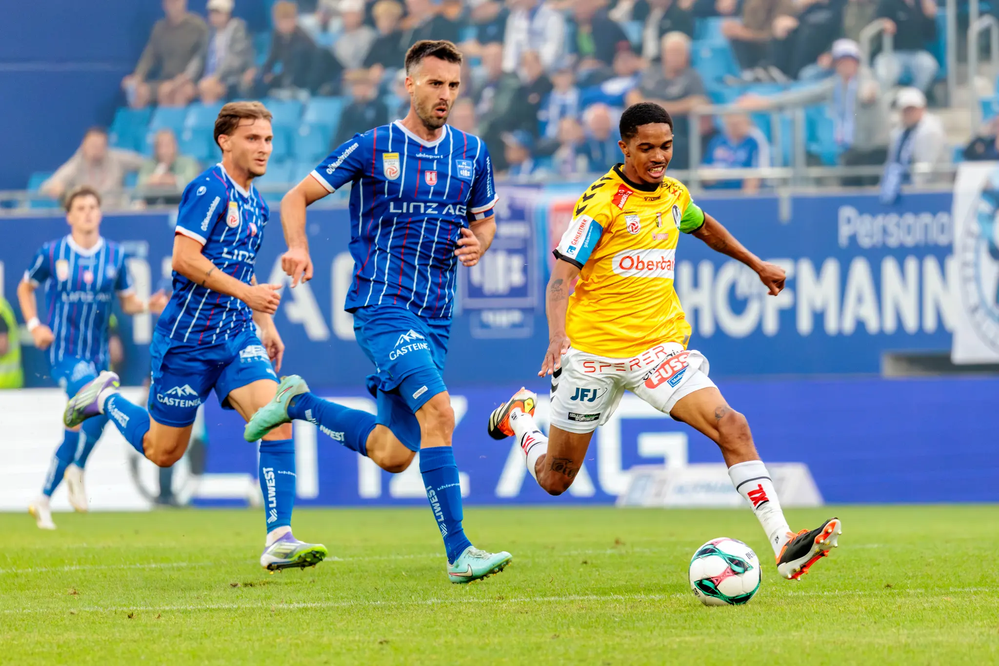 Ein Fußballspieler in gelbem Trikot dribbelt den Ball, während zwei blaue Kontrahenten auf dem Spielfeld neben ihm laufen
