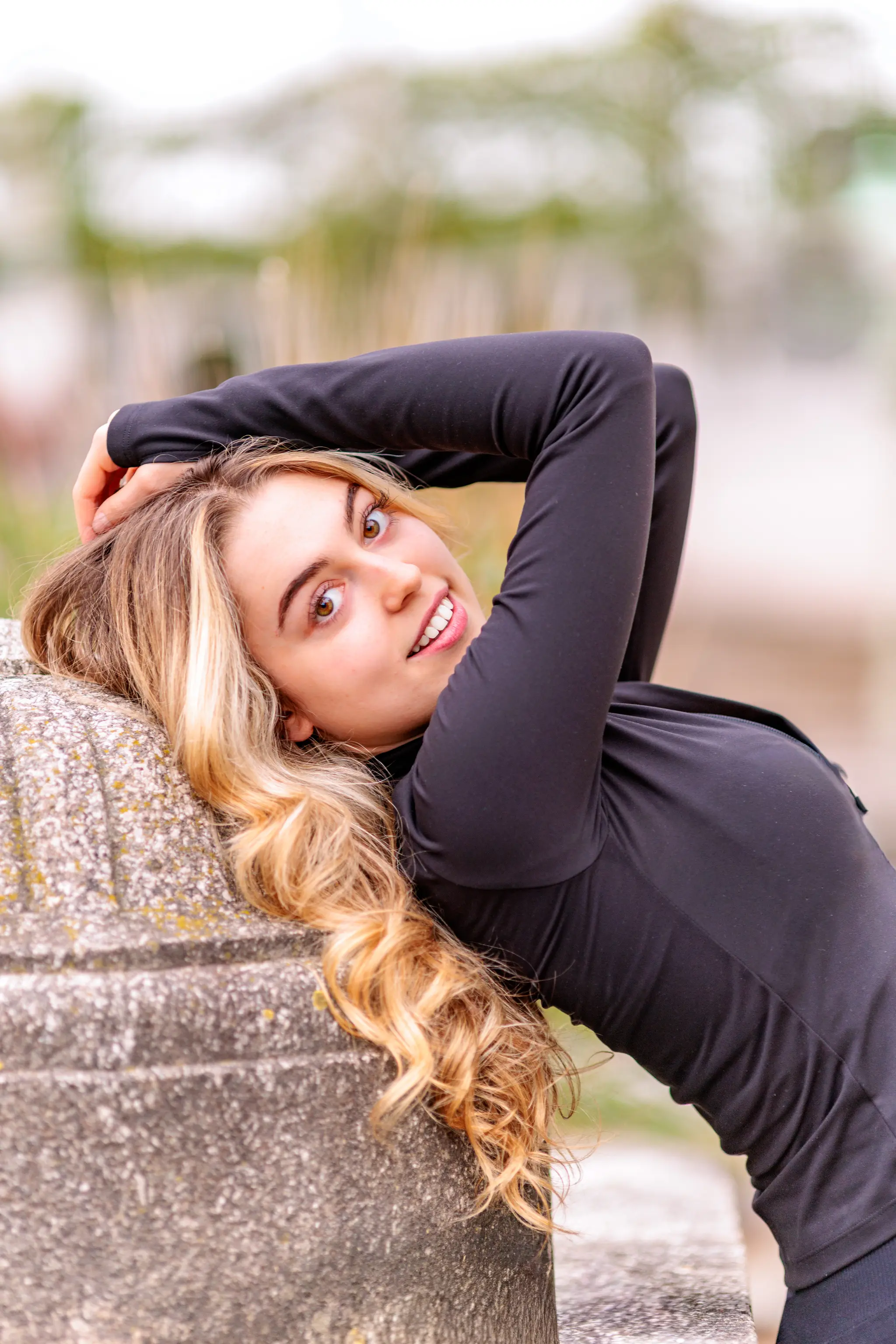 Das junge Model Valentina lehnt sich mit langen, blonden Haaren an eine Steinsäule, hat die Hände hinter dem Kopf und blickt lächelnd in die Kamera