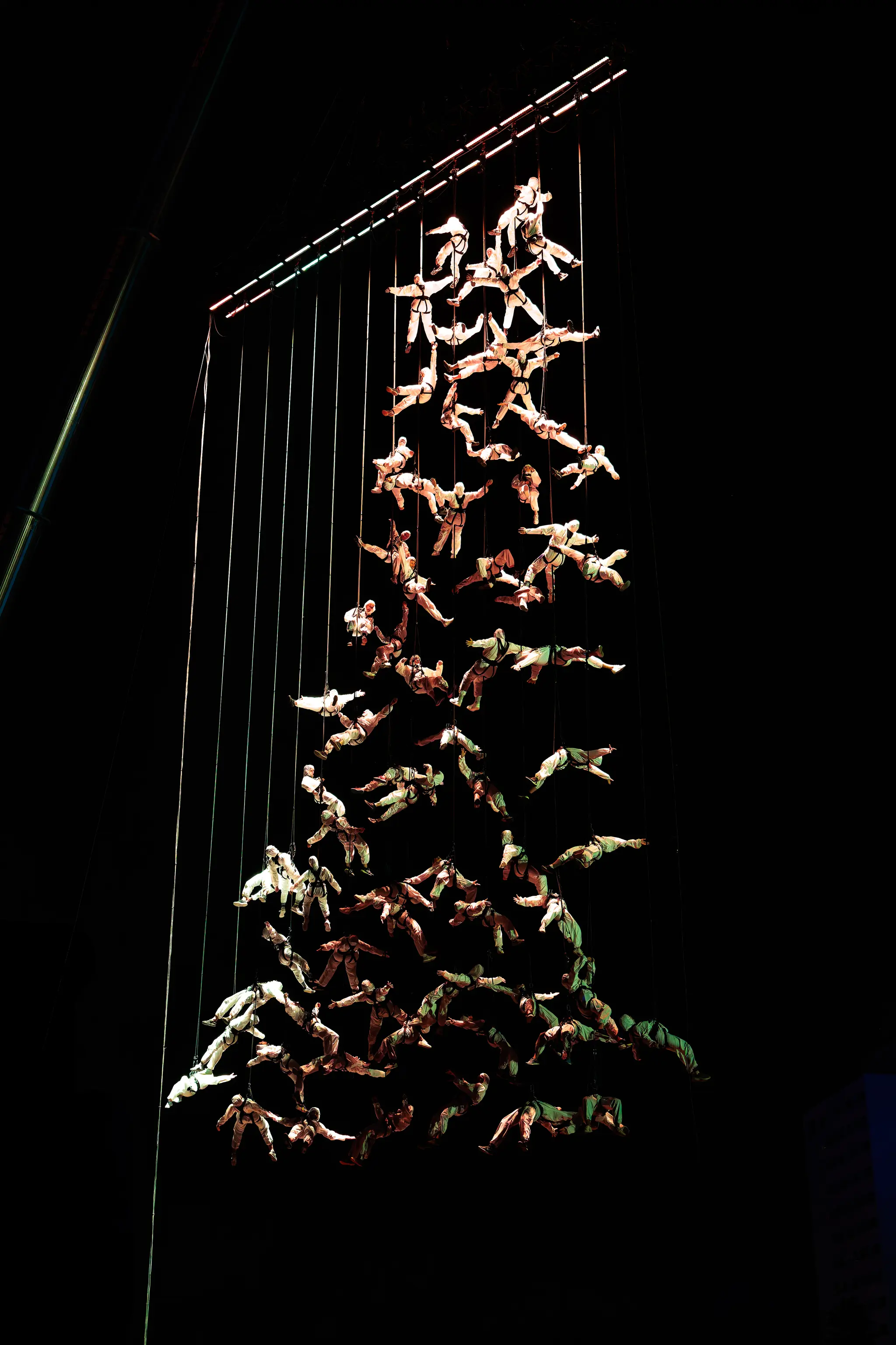 Luftakrobatik-Performance während der Linzer Klangwolke 2024 mit vielen Artisten in hellen Anzügen, die an Seilen von einer beleuchteten Deckenkonstruktion herabhängen und eine schwebende Formation vor dem nächtlichen Himmel bilden 