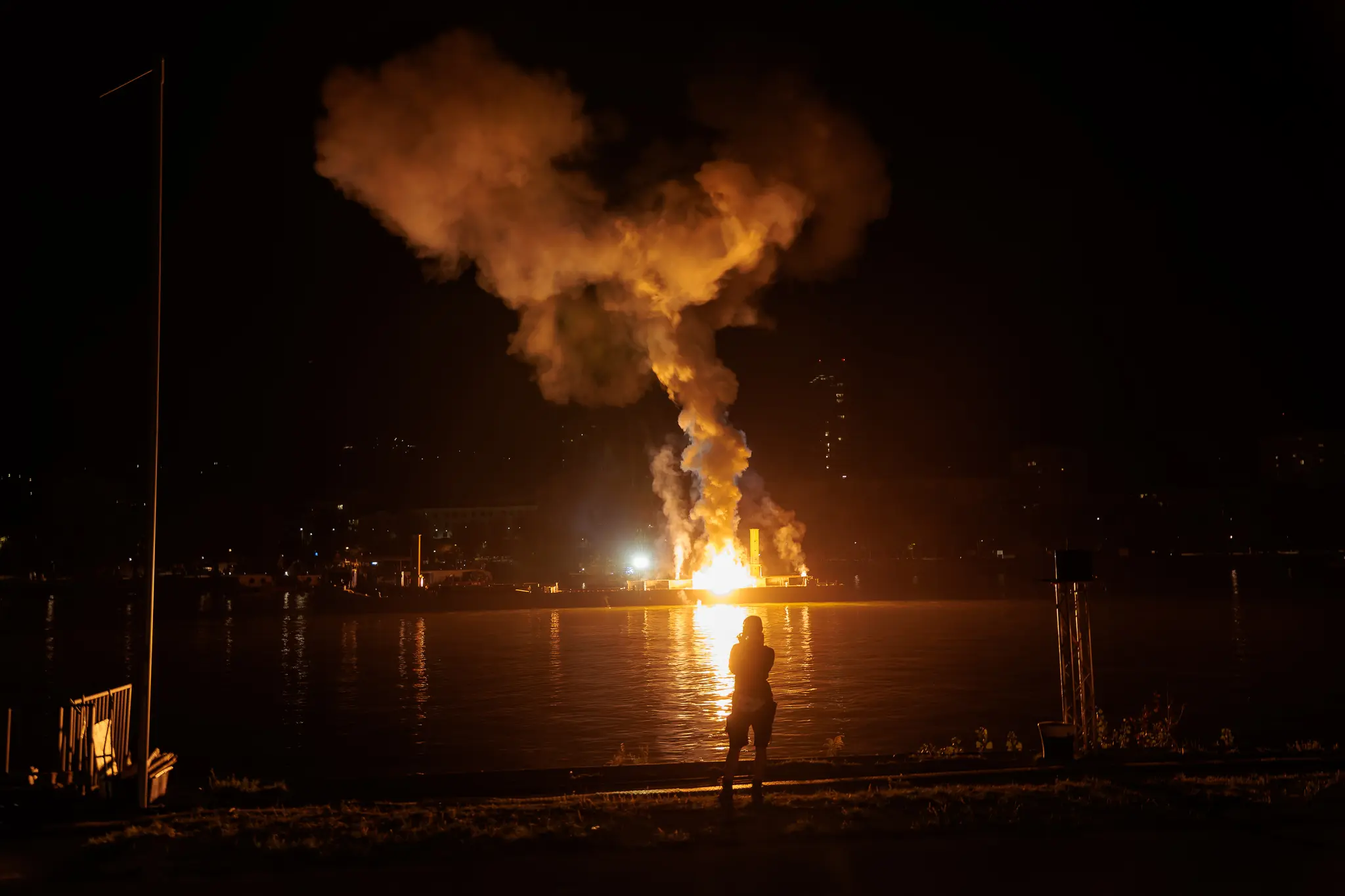 Nächtlicher Explosion mit orangefarbenem Rauch über der Donau als Abschluss der Klangwolke 2024 in Linz. Eine silhouettierte Person im Vordergrund beobachtet das Feuer 