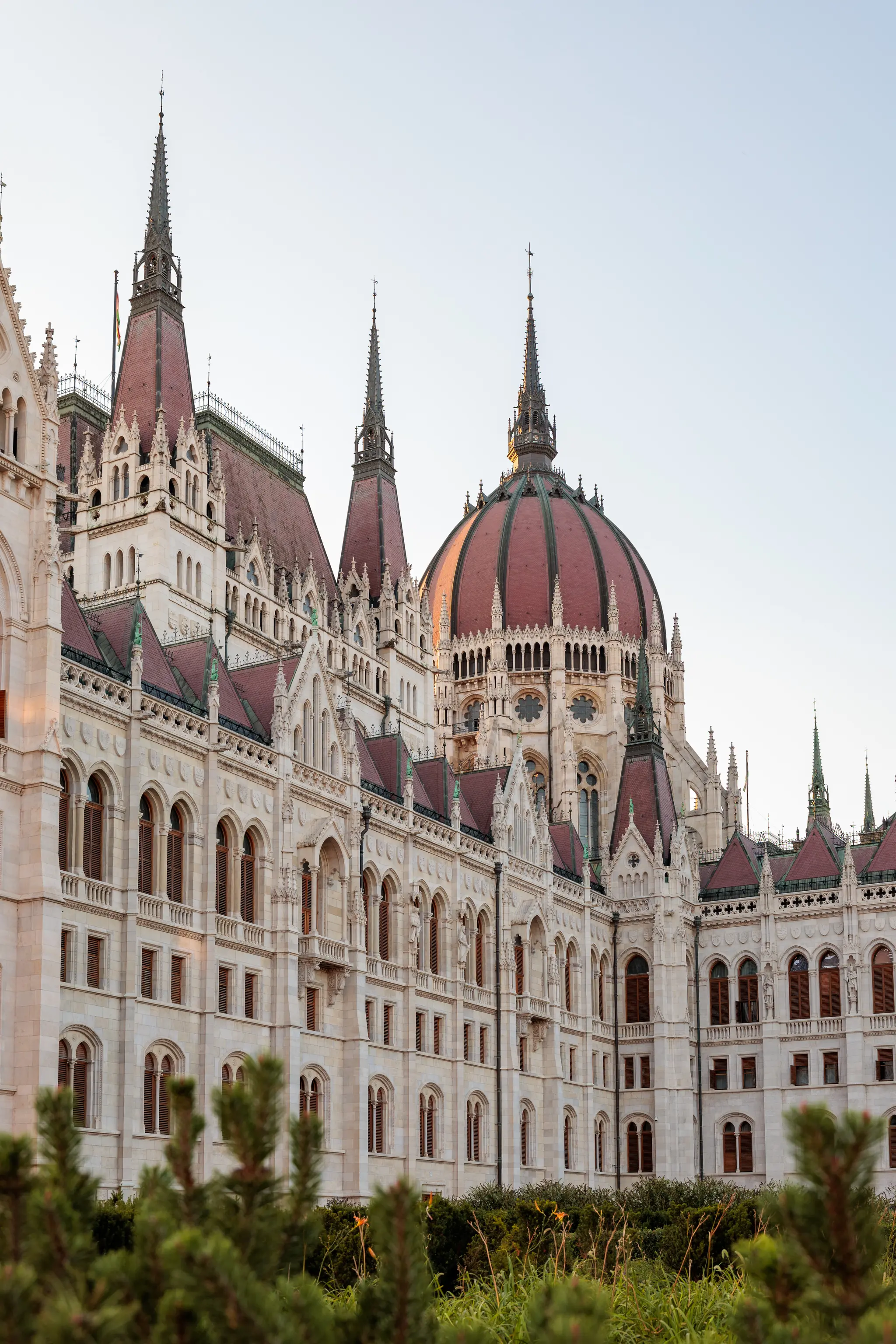 Das imposante Ungarische Parlament in Budapest, ein neugotisches Gebäude mit markanter Kuppel und zahlreichen Türmen, teilweise von Grünpflanzen im Vordergrund verdeckt. 