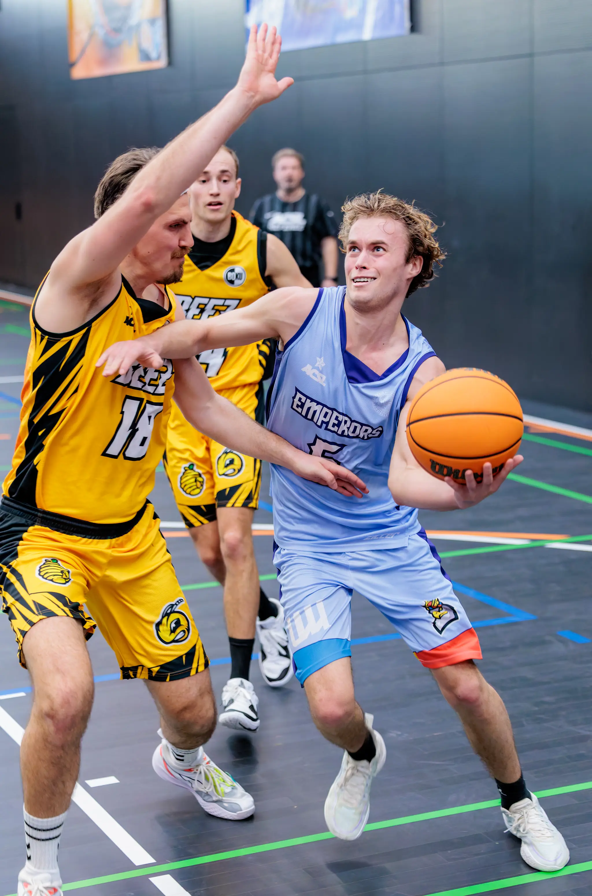 Ein Basketballspieler in hellblauem Trikot führt lächelnd den Ball, während ein gegnerischer Spieler in gelbem Trikot ihn defensiv herausfordert