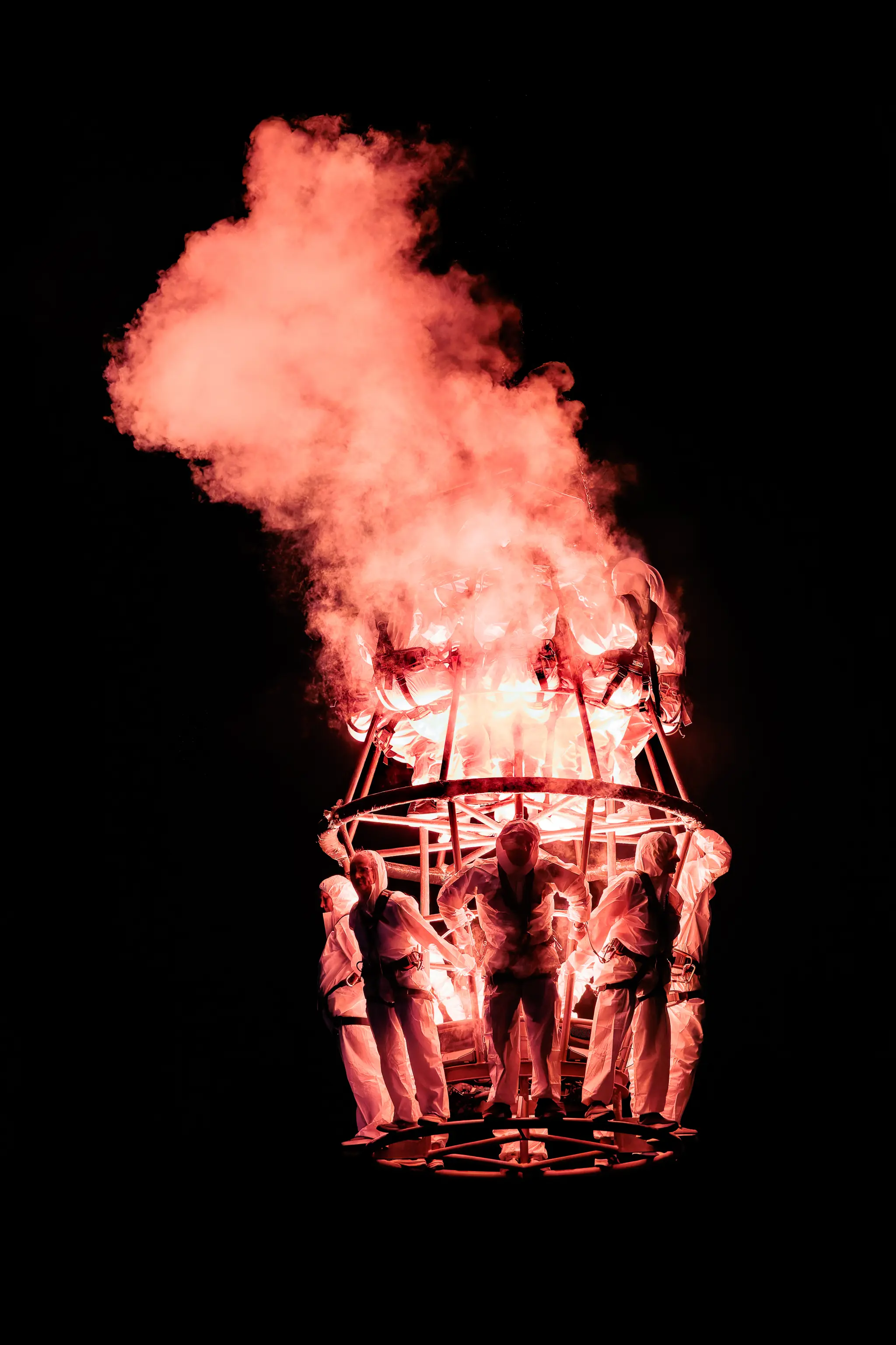 Dramatische Nachtszene einer brennenden Metallkonstruktion mit intensivem rotem Rauch und Menschen in weißen Schutzanzügen. Abstrakte Kunstperformance während des Pflasterspektakels 2024 in Linz 