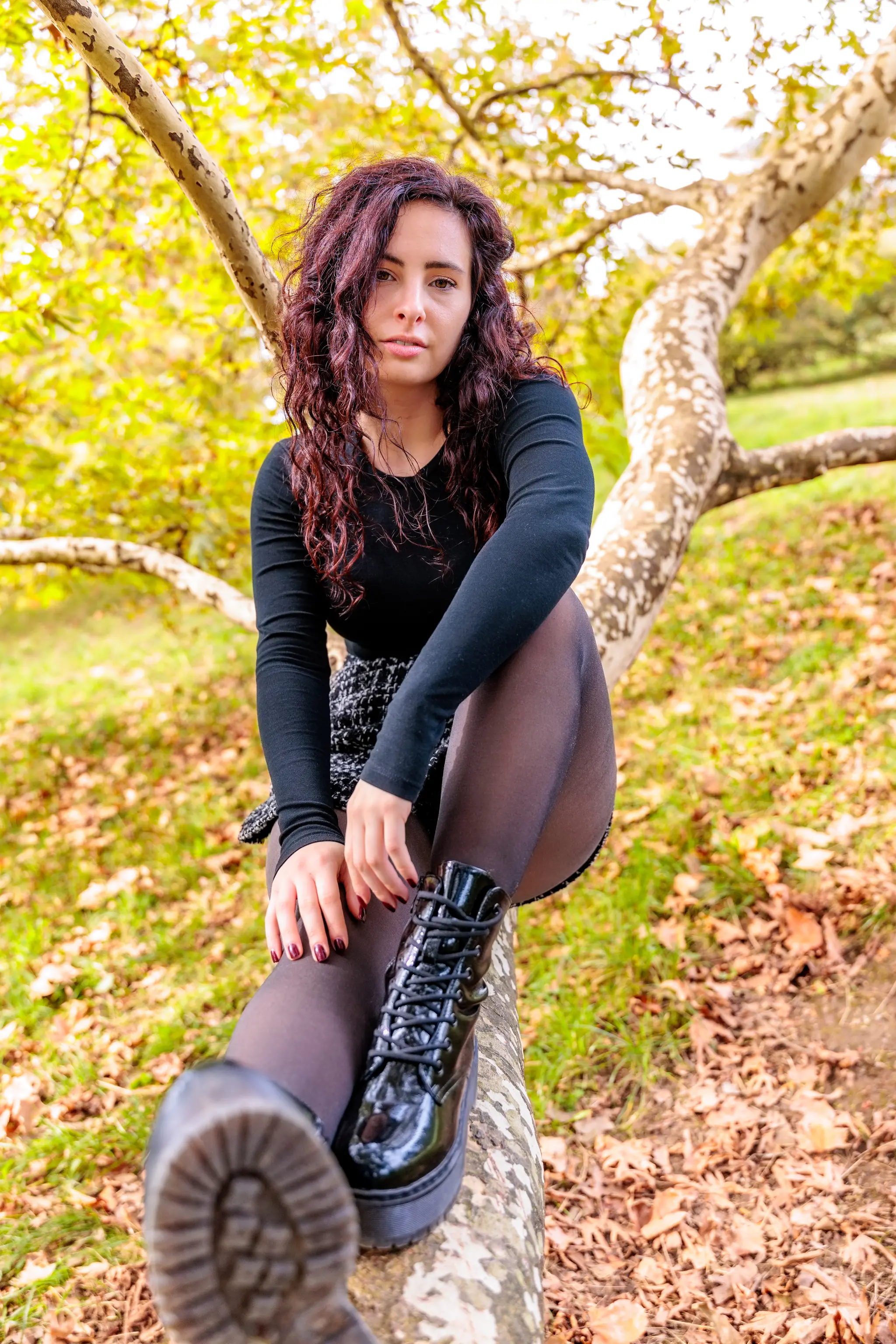 Junge Frau mit langen, dunkelroten Locken in schwarzem Oberteil, dunkler Strumpfhose und glänzenden schwarzen Plateau-Stiefeln sitzt auf einem markanten Baumstamm im Herbstlaub im Freien und blickt direkt in die Kamera.