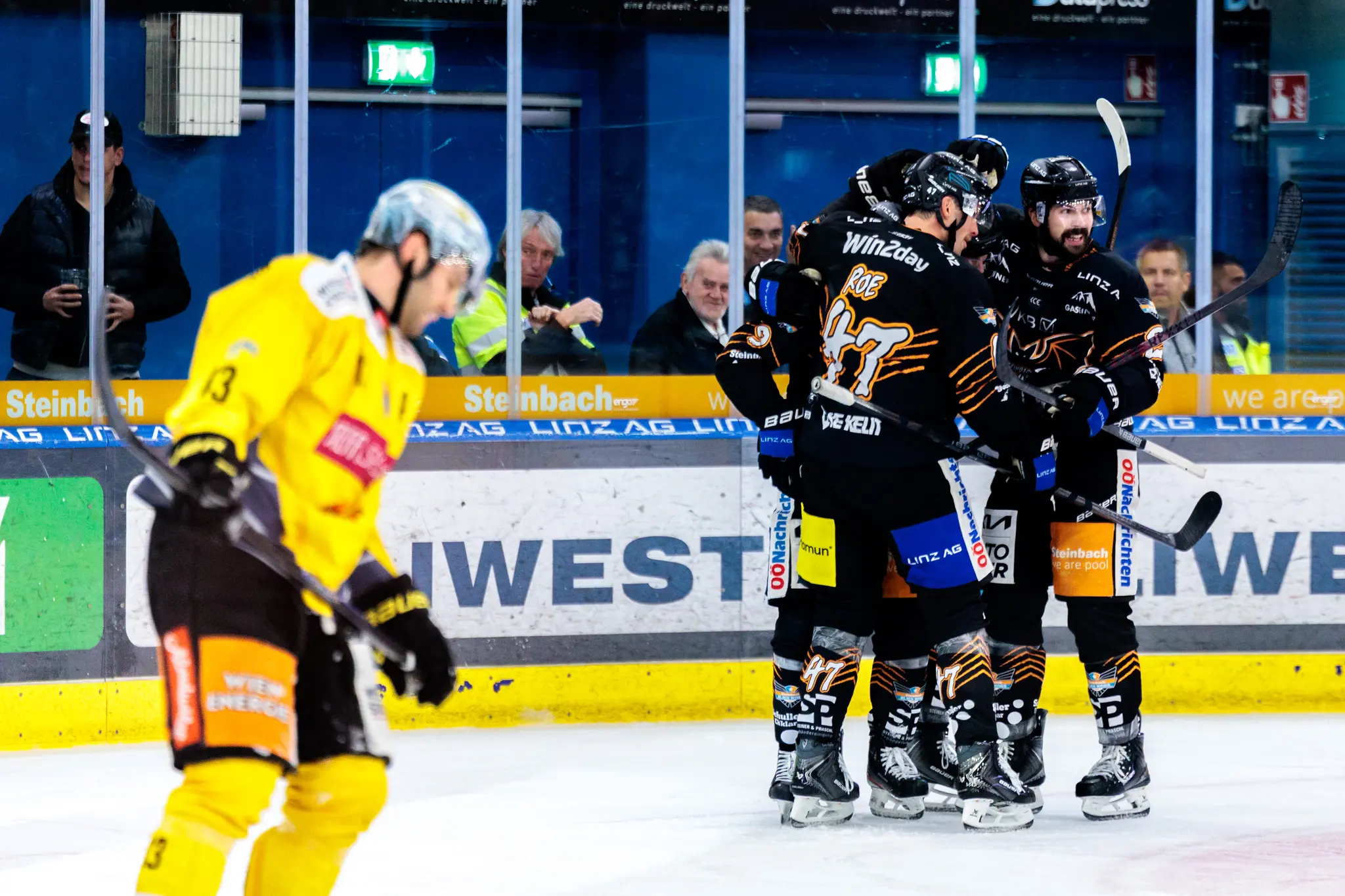 Die Eishockeyspieler der Black Wings Linz, angeführt von Logan Roe (#47), feiern einen Torerfolg auf dem Eis, während im Vordergrund ein enttäuschter Spieler der Vienna Capitals wegläuft