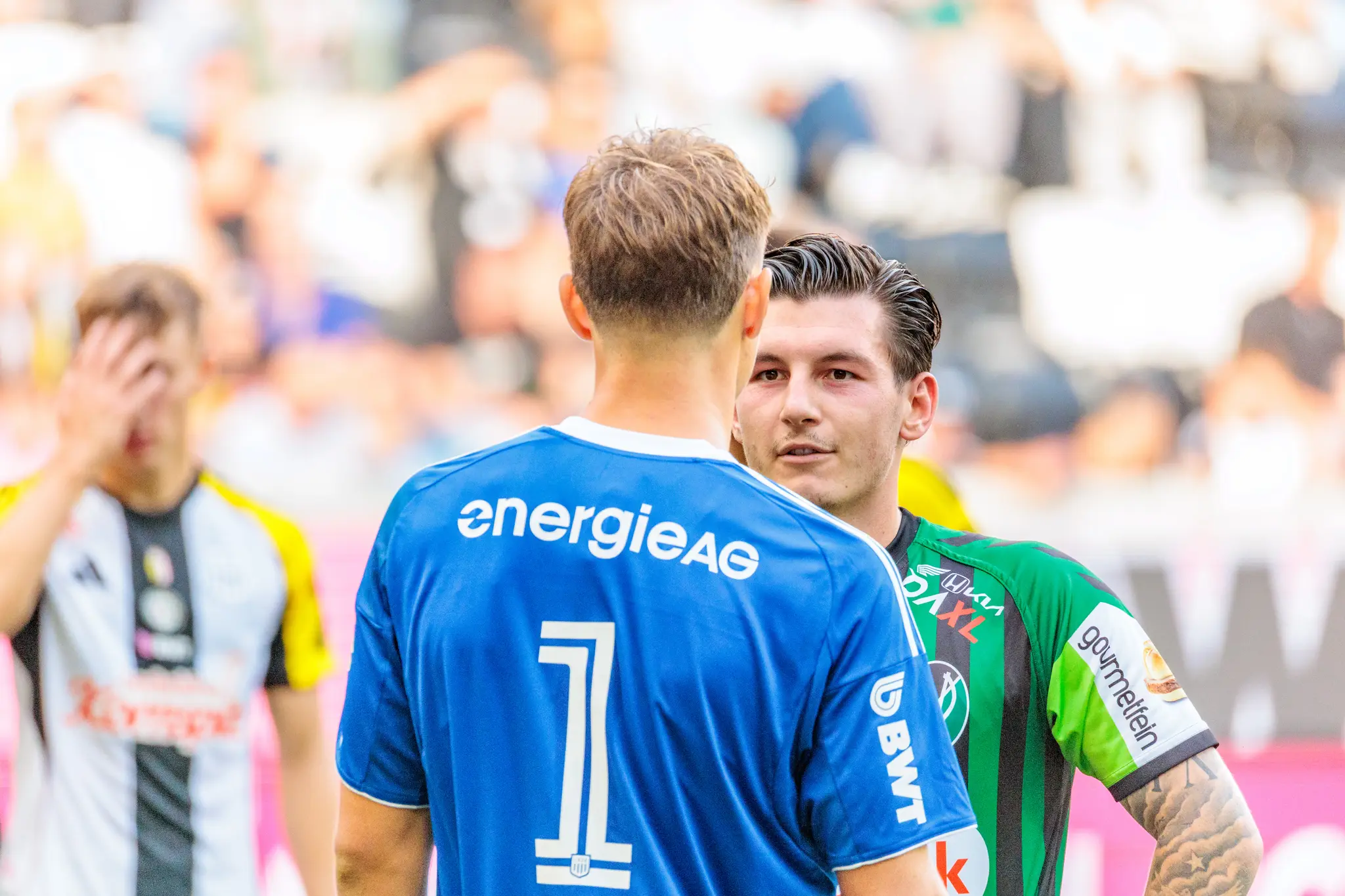 Ein LASK-Torhüter im blauen Trikot mit der Nummer 1 unterhält sich mit einem SV Ried-Spieler in grün-schwarzem Trikot auf dem Fußballfeld vor der Ausführung eines Elfmeters während eines Bundesliga-Spiels