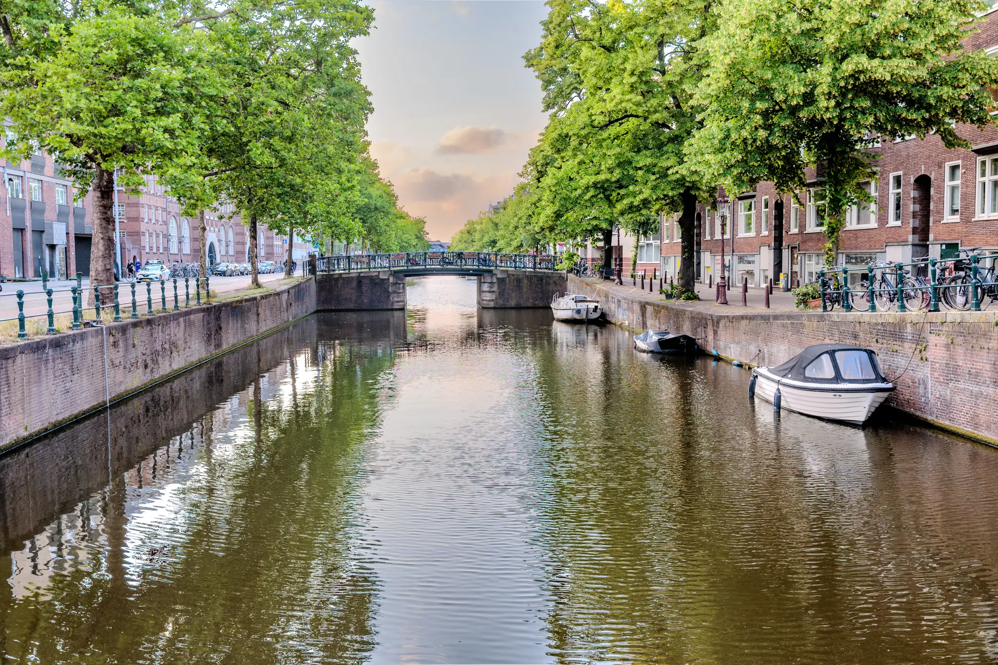 Ein malerischer Kanal in Amsterdam, gesäumt von üppigen Bäumen und traditionellen Gebäuden, mit Booten, die im Abendlicht ruhig auf dem Wasser liegen 