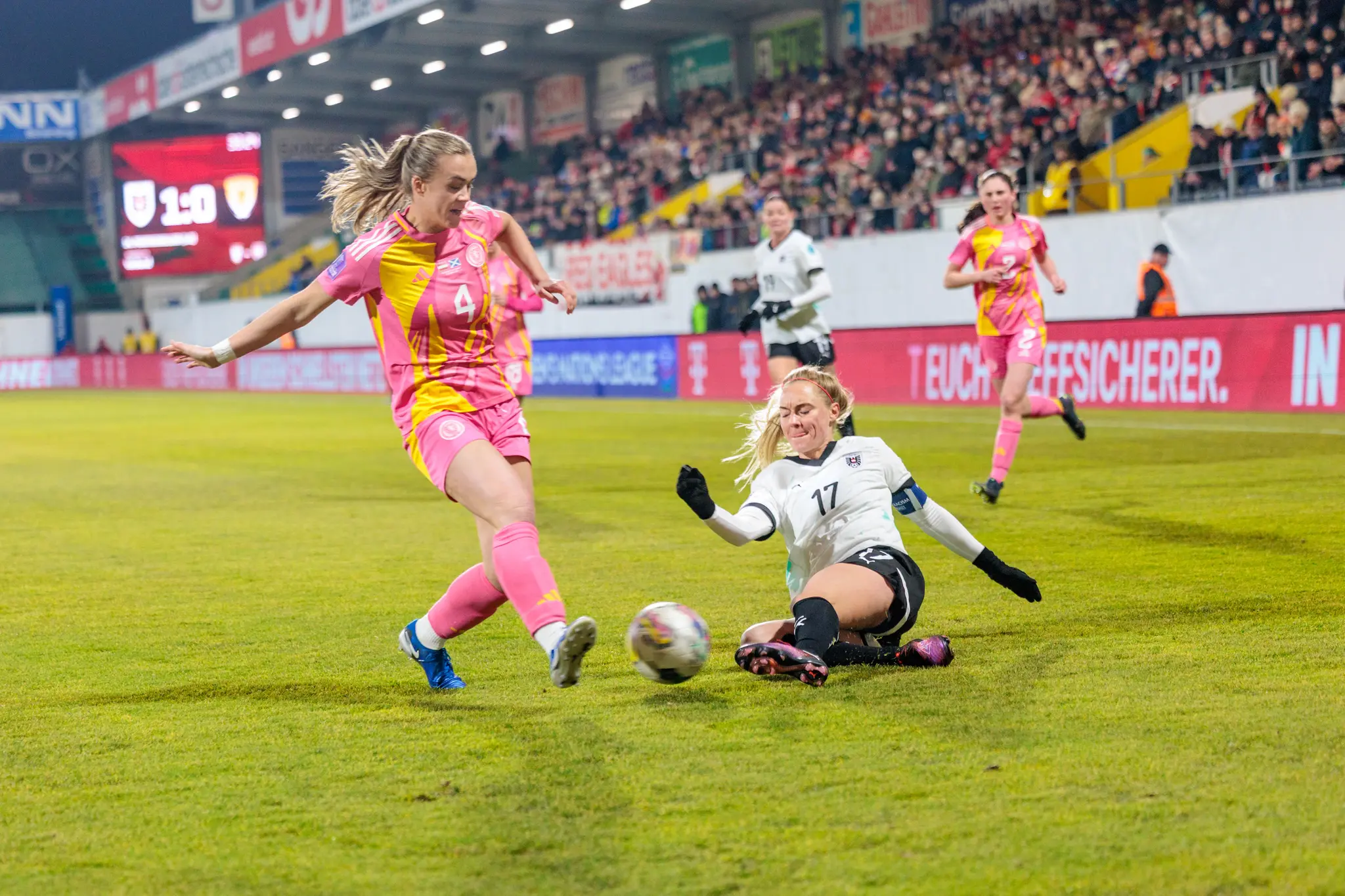Eine schottische Fußballerin in Pink und Gelb versucht den Schuss einer österreichische Spielerin in Weiß, die am Boden liegt, zu blockieren