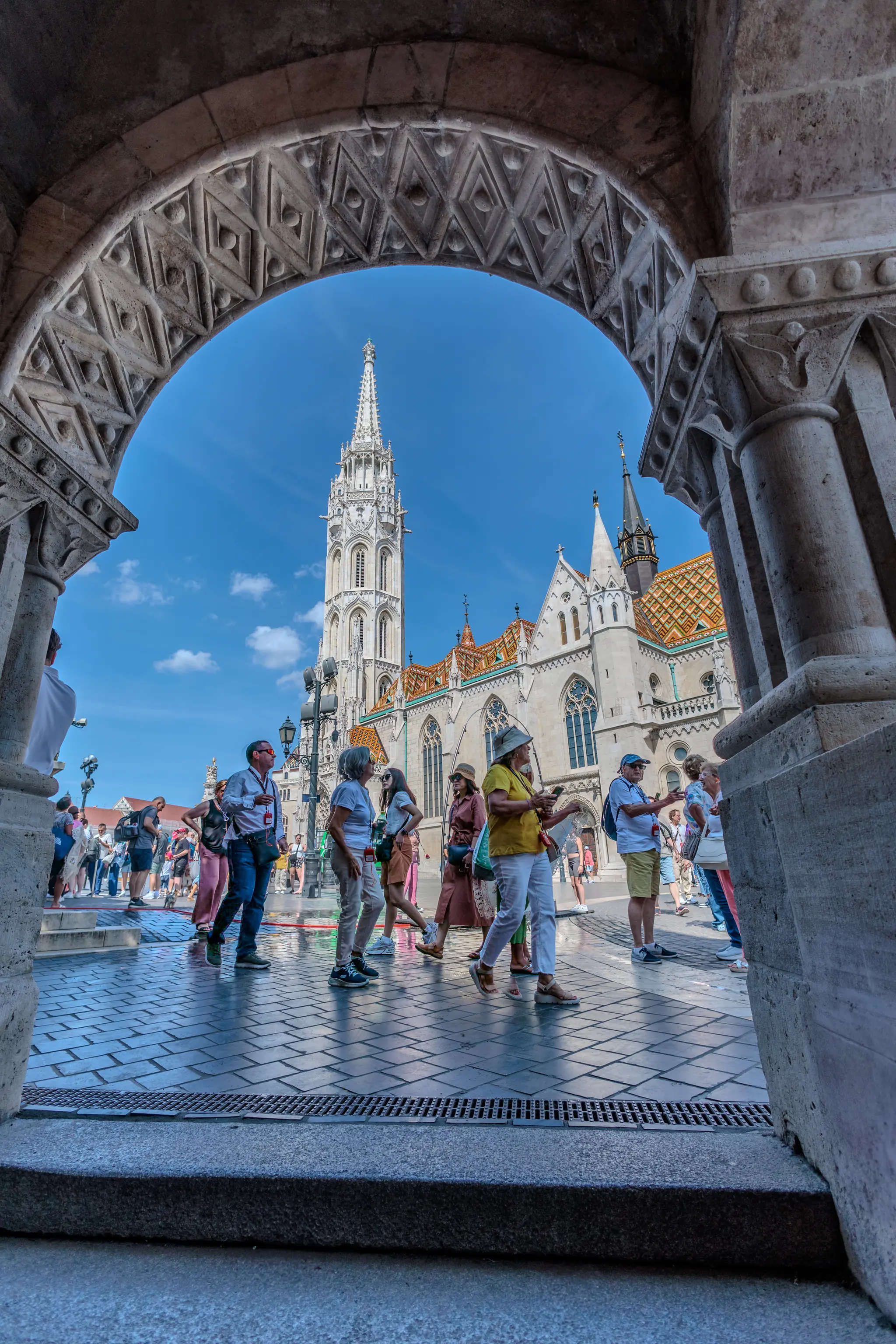 Die Matthiaskirche in Budapest ist durch einen verzierten Bogen der Fischerbastei zu sehen, mit Touristen auf dem belebten Platz unter blauem Himmel. 