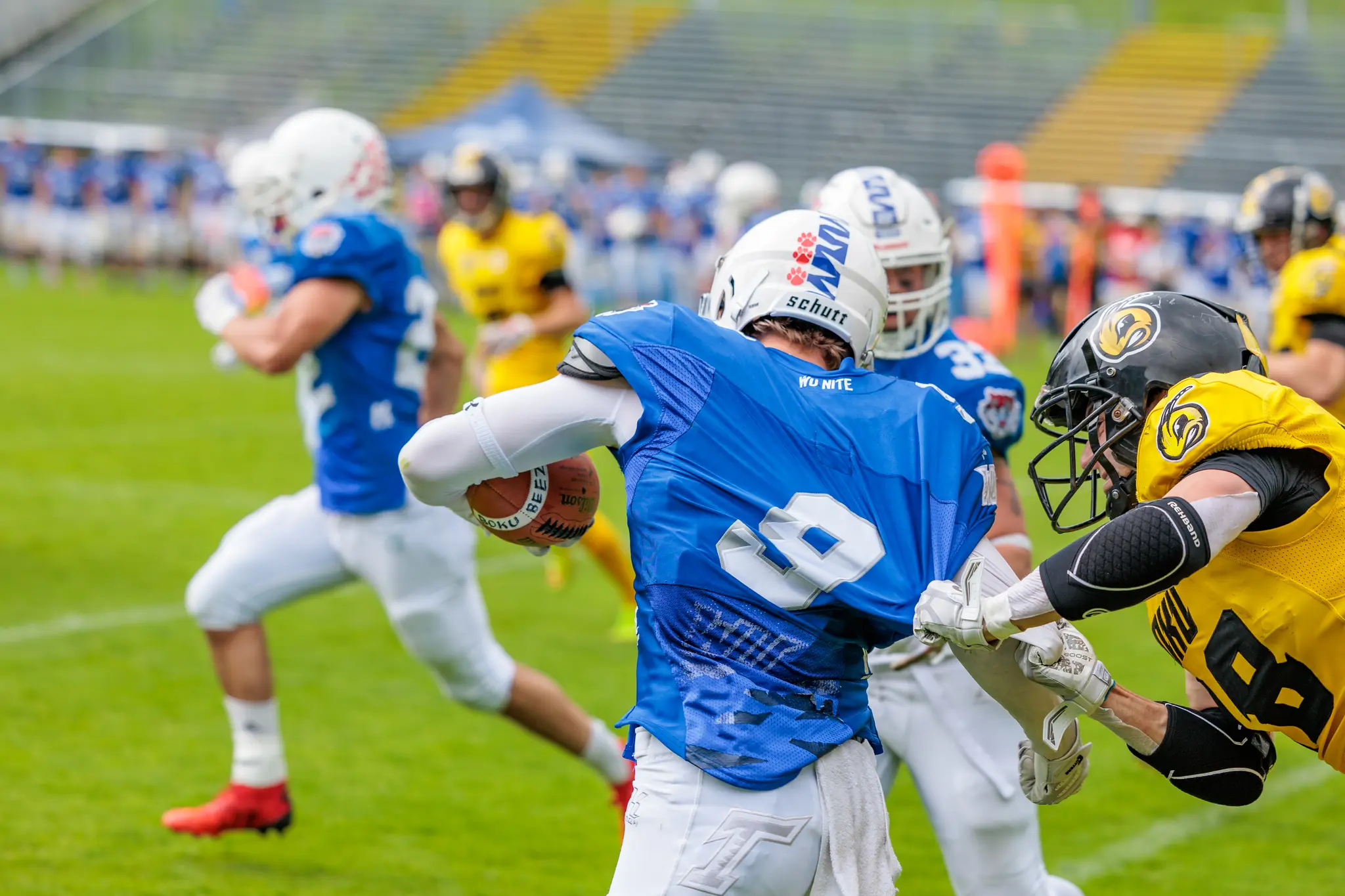 Ein Footballspieler in blauem Trikot versucht, einem Tackle zu entkommen, während ein gelb gekleideter Gegner sein Trikot packt