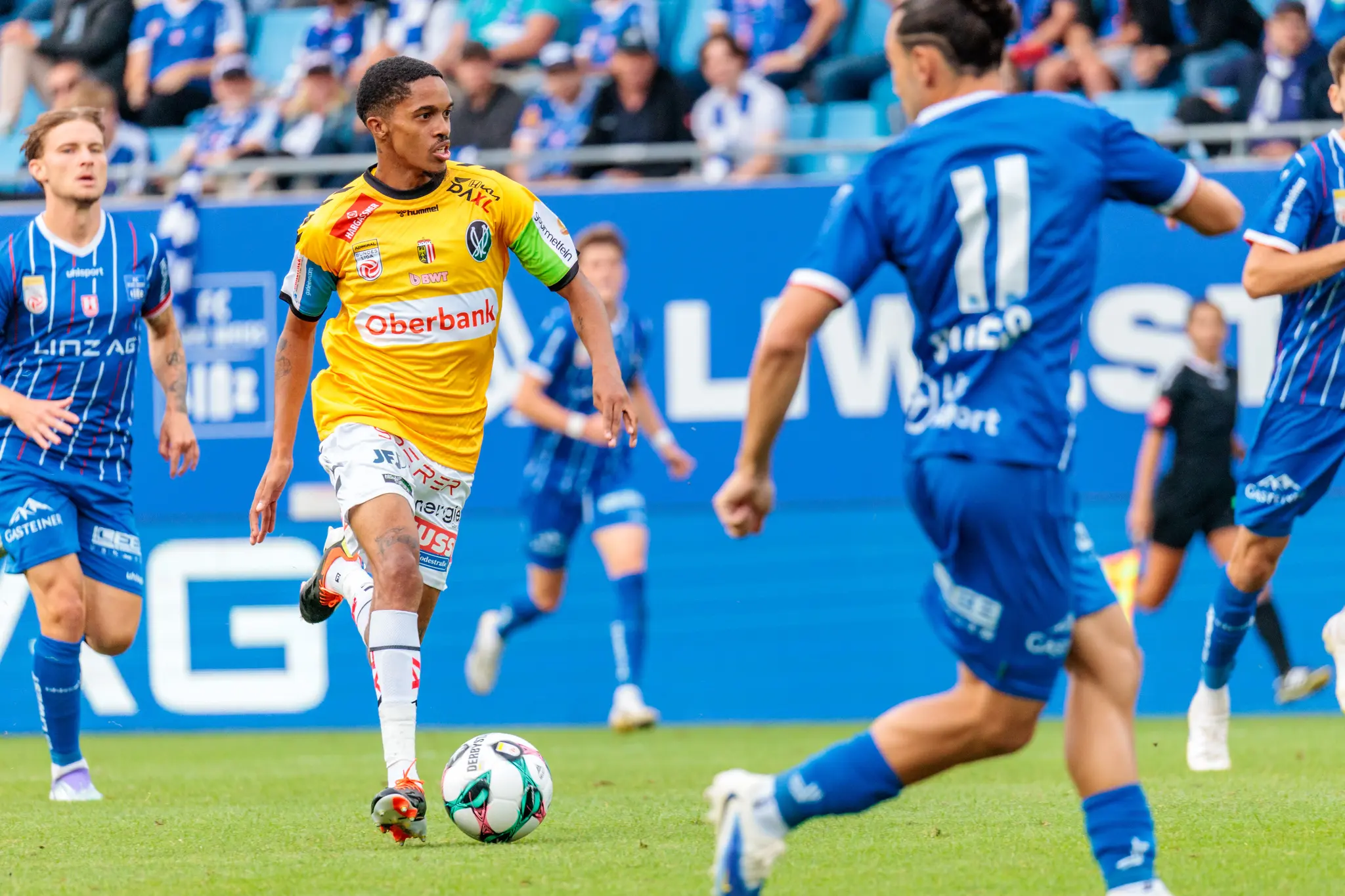 Ein Fußballspieler des SV Ried in gelbem Trikot dribbelt den Ball in einem intensiven Zweikampf gegen Spieler von Blau-Weiß Linz