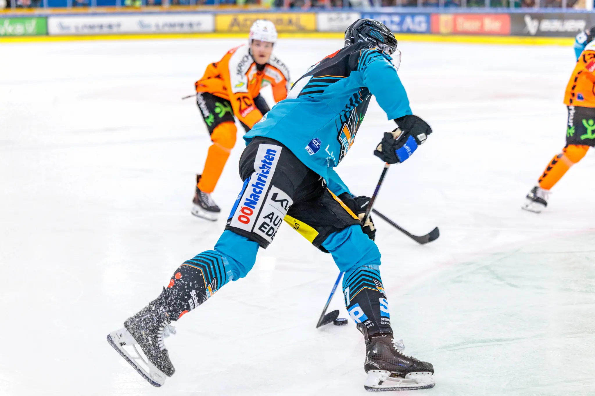 Ein Eishockeyspieler in türkisfarbenem und schwarzem Trikot gleitet über das Eis und erzeugt Eisstaub, während er in einem Zweikampf mit einem orange gekleideten Gegner ist