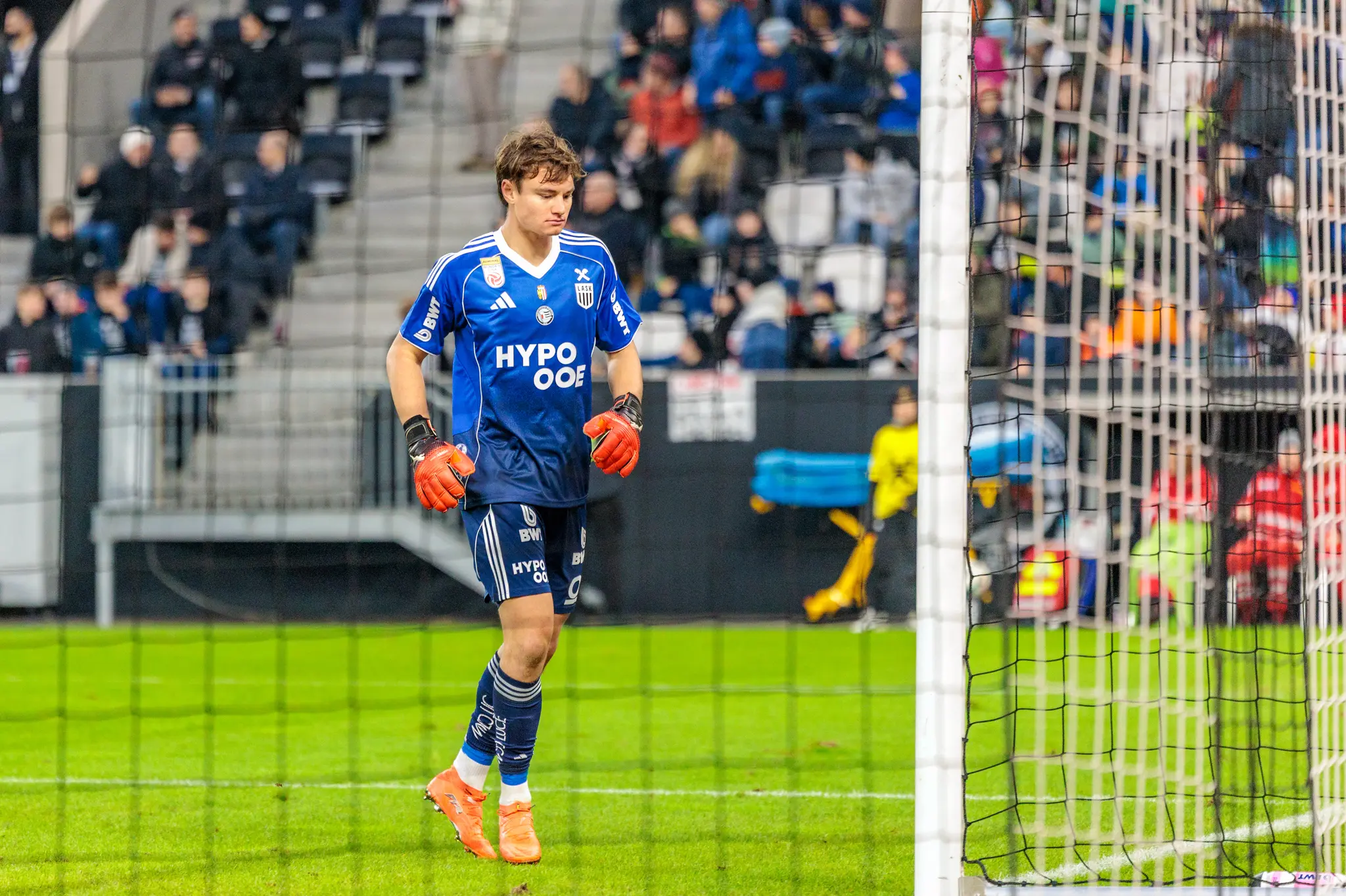 Ein junger Fußballtorwart des LASK im blauen Trikot mit orangefarbenen Handschuhen und Schuhen steht konzentriert vor einem Tornetz