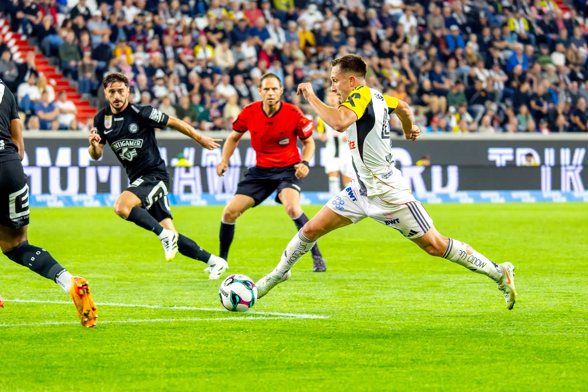 Dynamisches Bild eines Fußballspielers des LASK, der gerade einen Ball tritt, umgeben von einem Schiedsrichter, einem Gegenspieler und Zuschauern in der Raiffeisen Arena