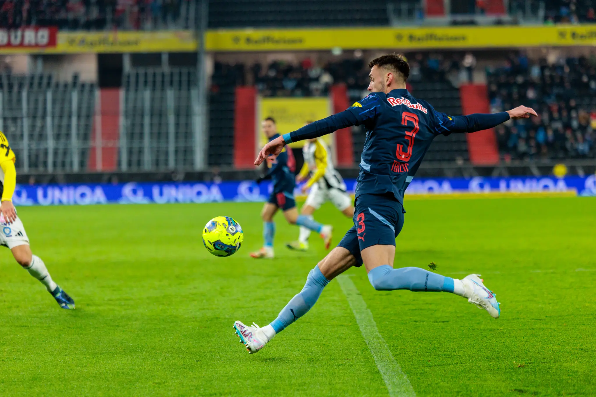 Dynamische Fußballszene: Spieler in Red Bull Trikot (Nr. 3) tritt gelb-grünen Ball auf grünem Stadionrasen 