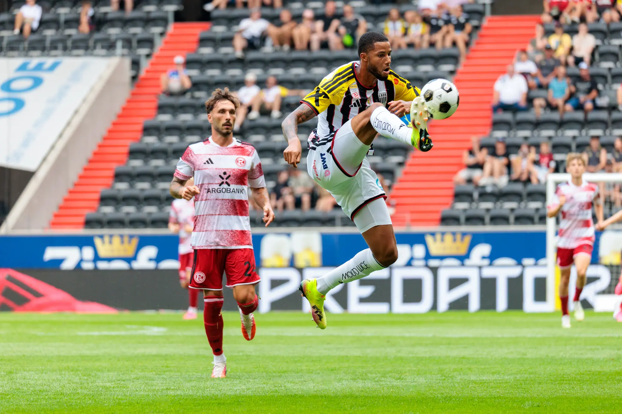 LASK-Spieler springt akrobatisch zum Ball und kontrolliert ihn mit dem Oberschenkel in der Luft, ein Düsseldorfer Spieler beobachtet die Aktion.
