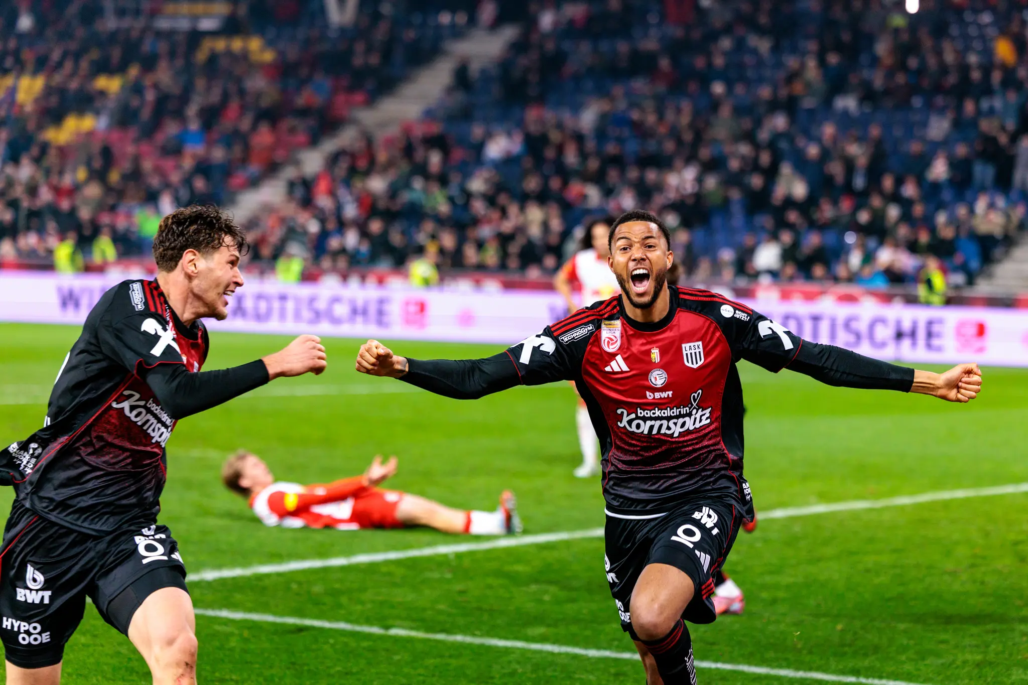 Zwei jubelnde LASK-Spieler feiern ekstatisch einen späten Siegtreffer im Bundesligaspiel gegen Red Bull Salzburg, während ein Gegenspieler enttäuscht am Boden liegt