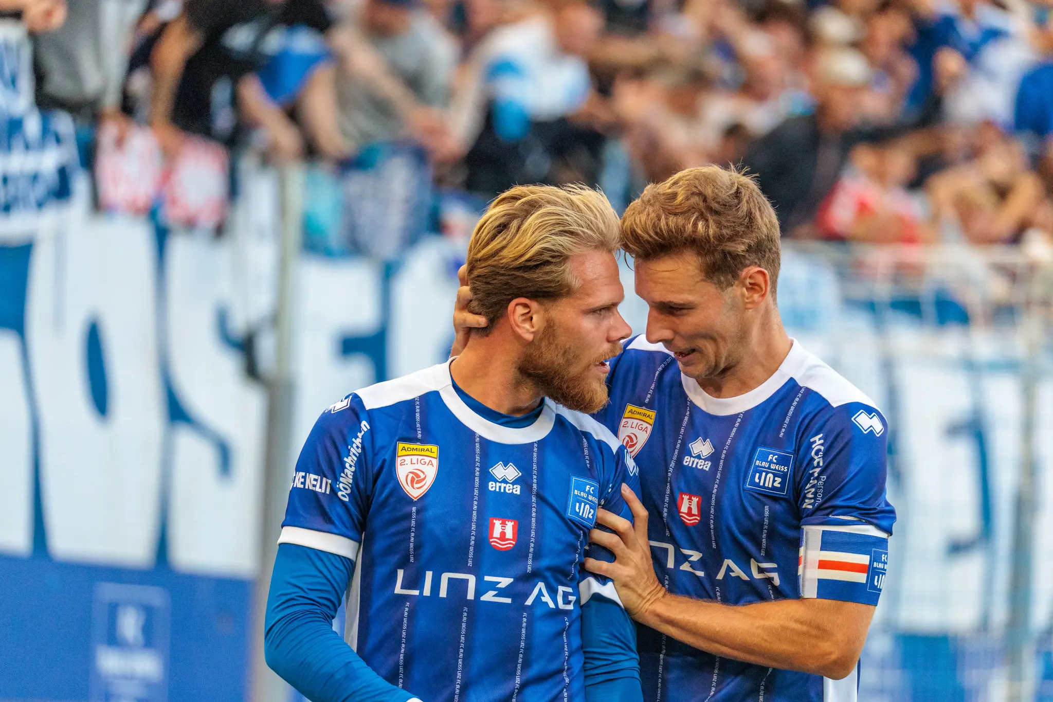 Zwei Linzer Spieler feiern ein Tor: Der Kapitän legt dem Teamkollegen die Hand an den Kopf und zieht ihn zu sich, beide lachen; im Hintergrund ist die blau-weiße Heimtribüne zu sehen.