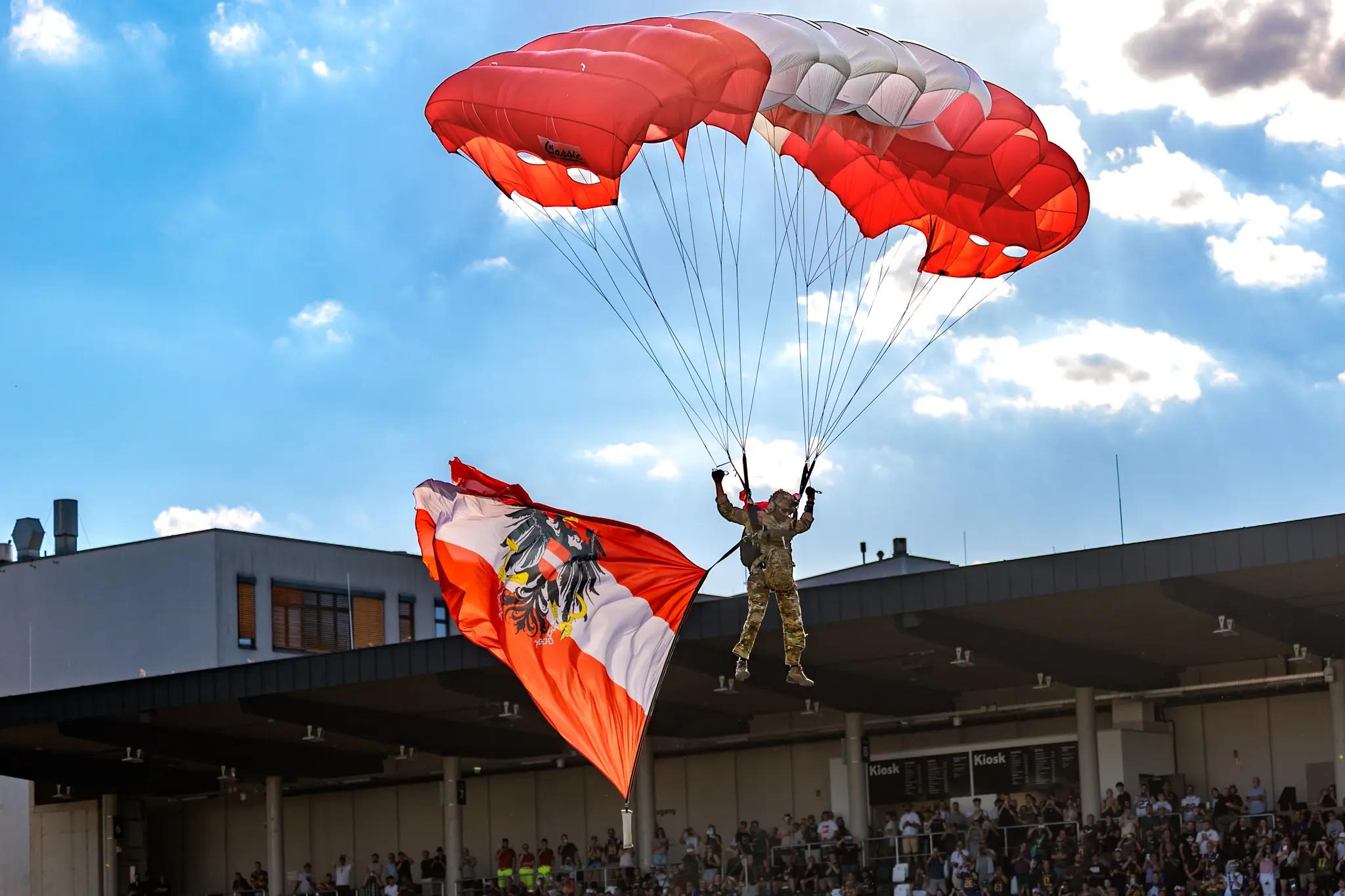 Ein Bundesheer-Springer schwebt mit rot-weißem Fallschirm und österreichischer Flagge über dem Tribünenbereich.