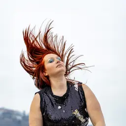 Porträt einer Frau mit hochgeworfenen roten Haaren und blauem Lidschatten in einem schwarzen Paillettenoberteil vor hellem Himmel