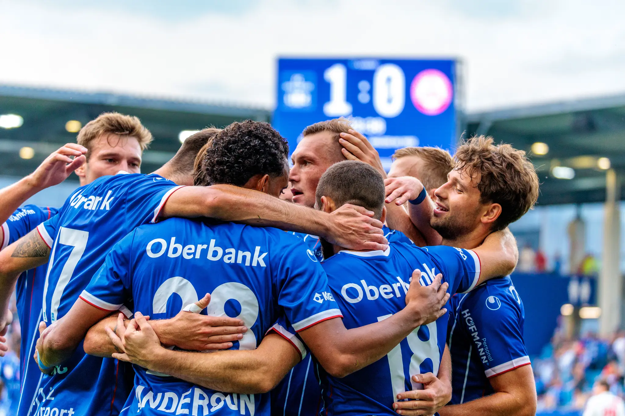 Blau-Weiß Linz Fußballspieler in blauen Trikots feiern eng umschlungen einen Führungstreffer, während die Anzeigetafel im Hintergrund das Ergebnis 1:0 anzeigt
